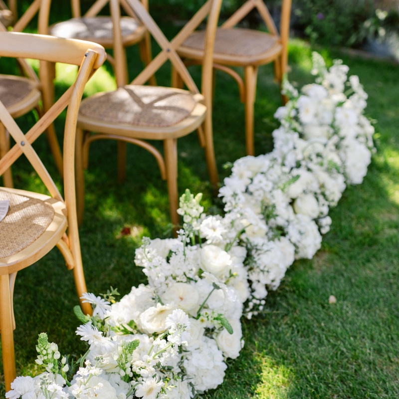 Allée de cérémonie de mariage extérieure bordée d'un tapis de fleurs blanches luxuriantes, devant des chaises de style crossback en bois.