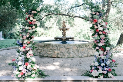 Allée de mariage extérieure ornée de deux piliers floraux richement décorés de roses et d'hortensias, menant à une fontaine en pierre.