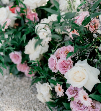 Arrangement floral luxuriant au sol avec des roses blanches et roses, des gerberas pâles et un feuillage vert dense, posé sur des graviers clairs.