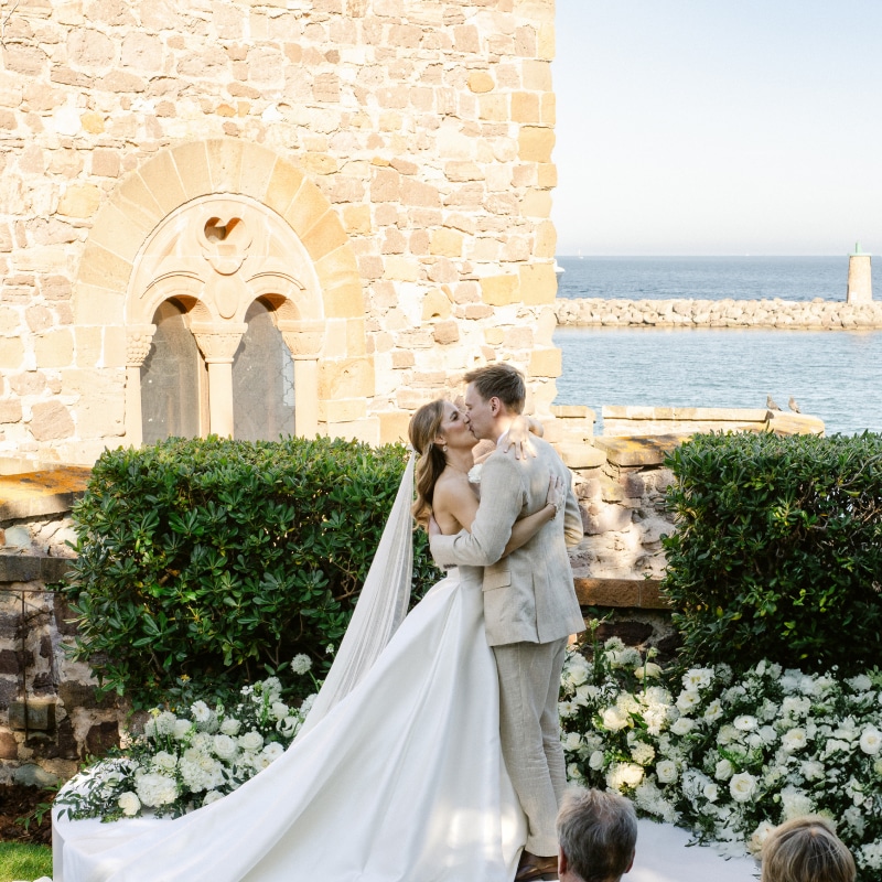 Jeunes mariés s'embrassant lors d'une cérémonie extérieure, avec un mur en pierre ancienne, une haie verte et la mer en arrière-plan.