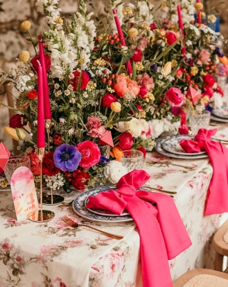 Table dressée avec centre de table floral riche et coloré, bougies fuchsia et linge à motif rose. Carte 'Un été qui l'est' visible.