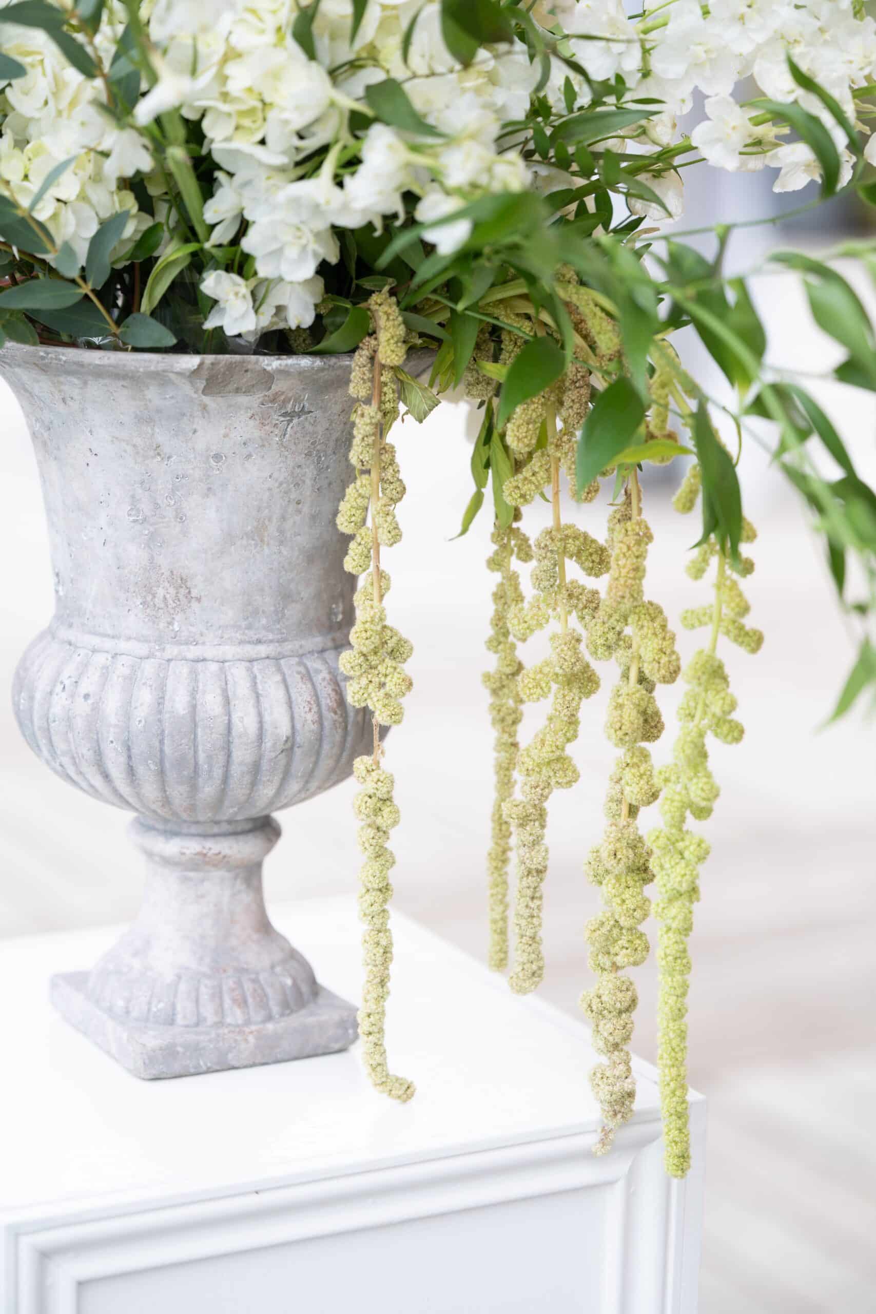 Urne grise patinée remplie de fleurs blanches. De longues amarantes vert pâle retombent élégamment devant le vase sur une console.