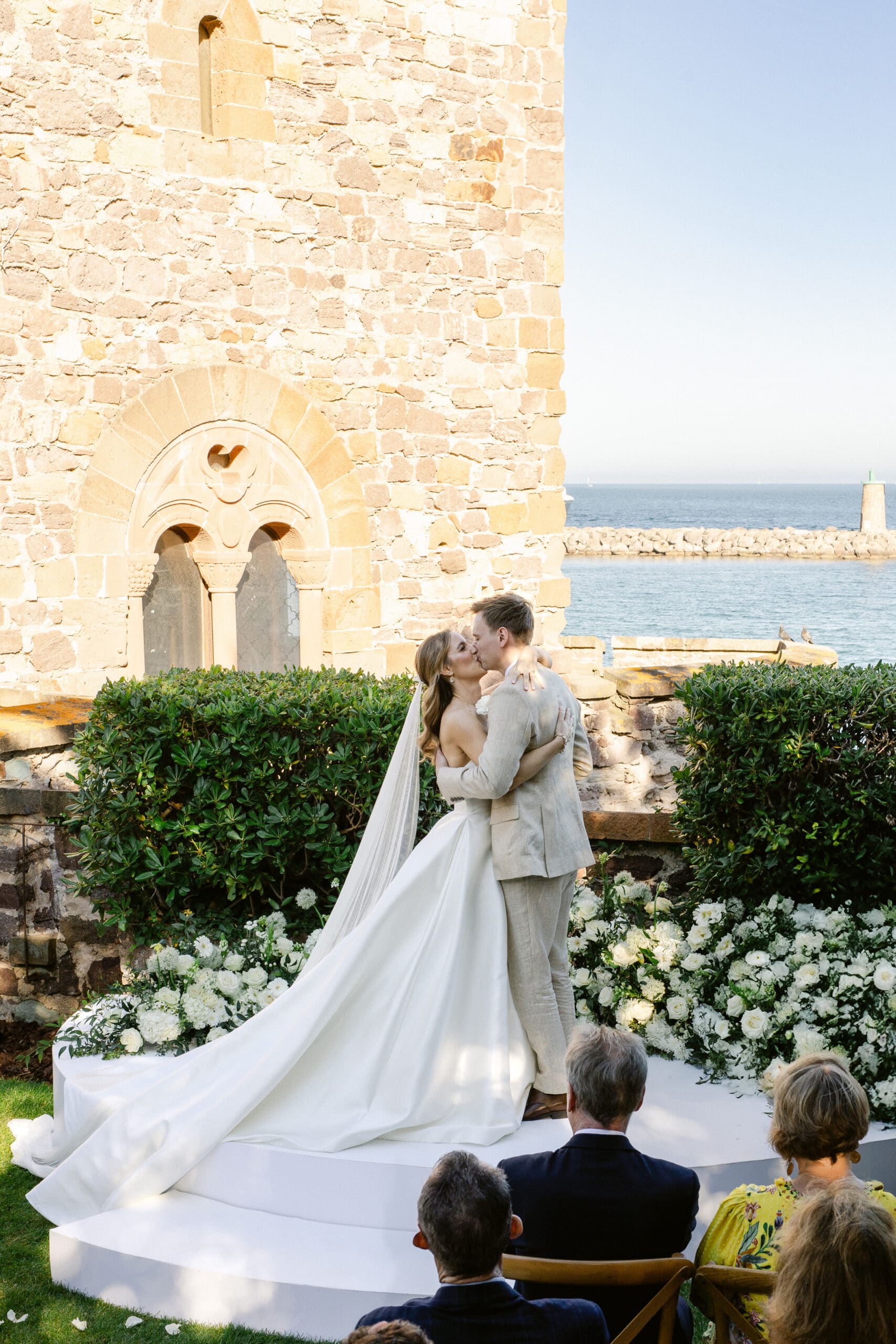 Baisé romantique des mariés sur une estrade. Robe de mariée en satin et costume lin clair, devant un mur en pierres anciennes et la mer Méditerranée.