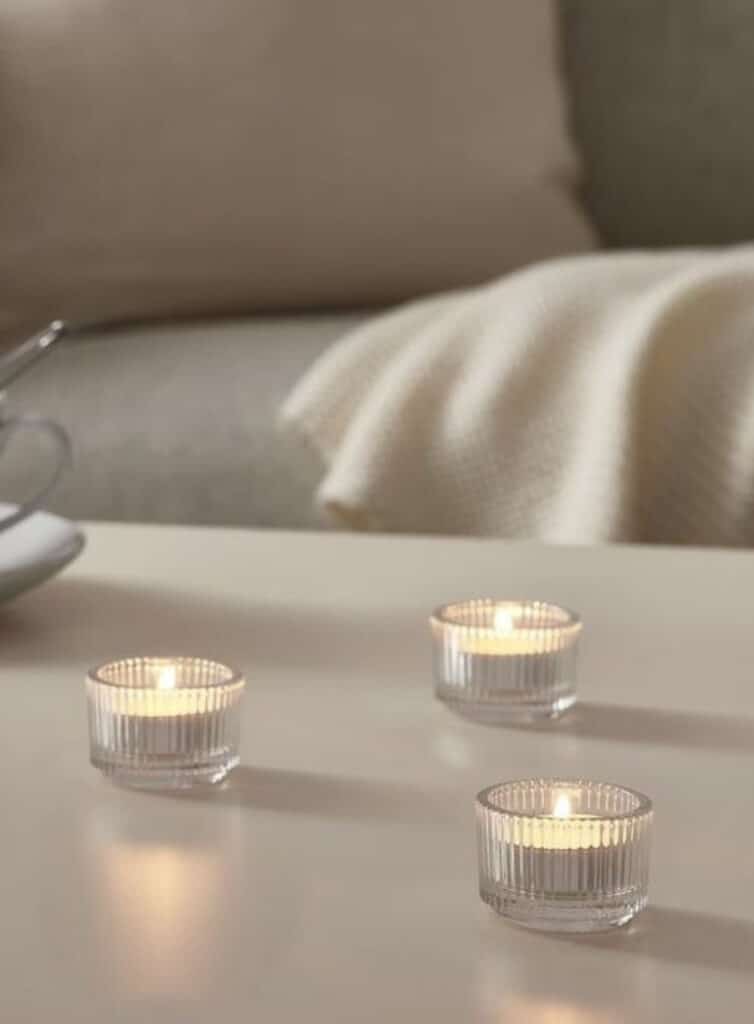 Ambiance chaleureuse. Trois bougies chauffe-plat allumées dans des photophores en verre strié sur une table, devant un canapé douillet avec un plaid.