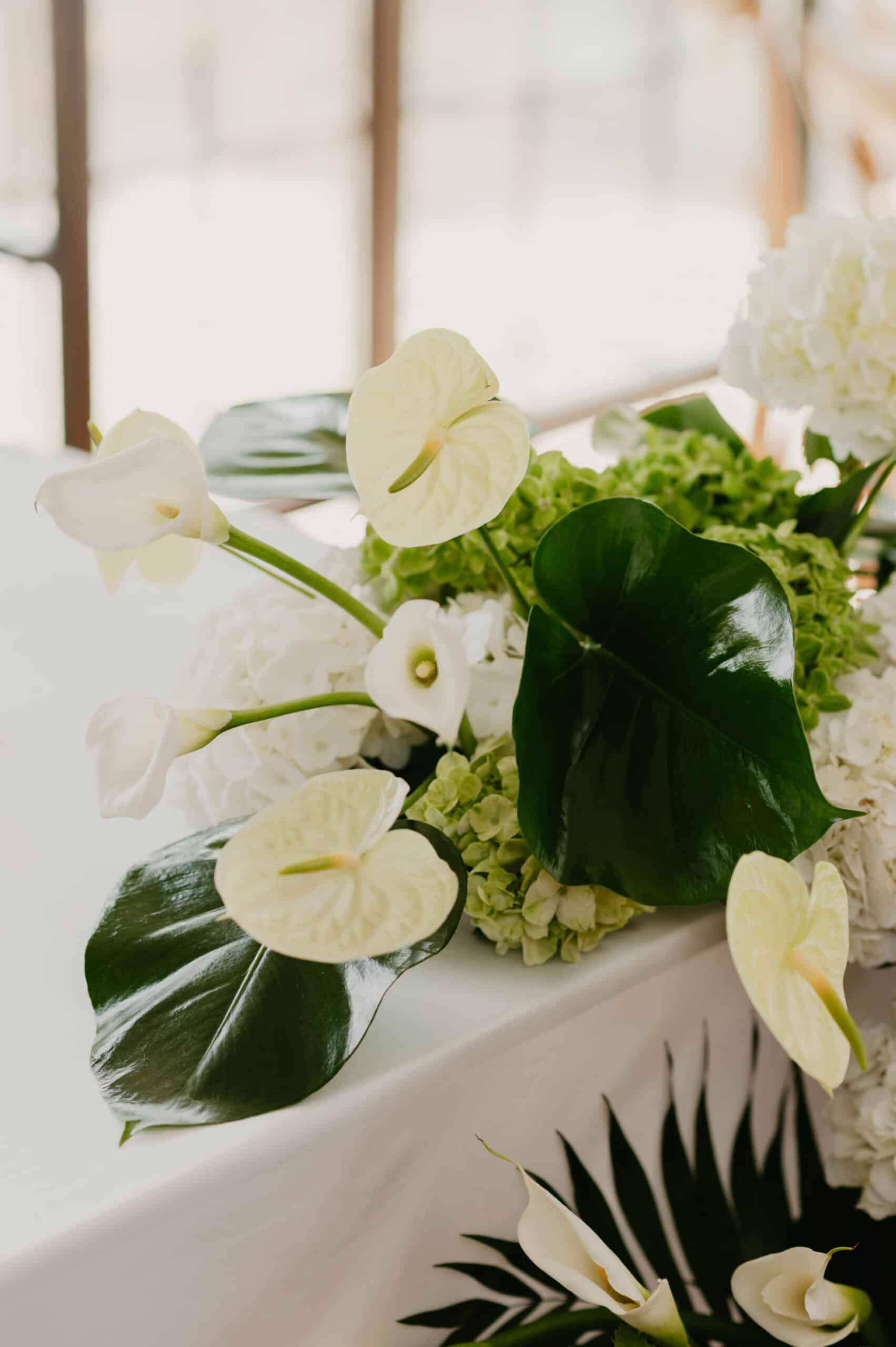 Composition florale élégante pour mariage. Calla blancs, Anthuriums crème, et Hydrangeas vert lime avec grandes feuilles tropicales.