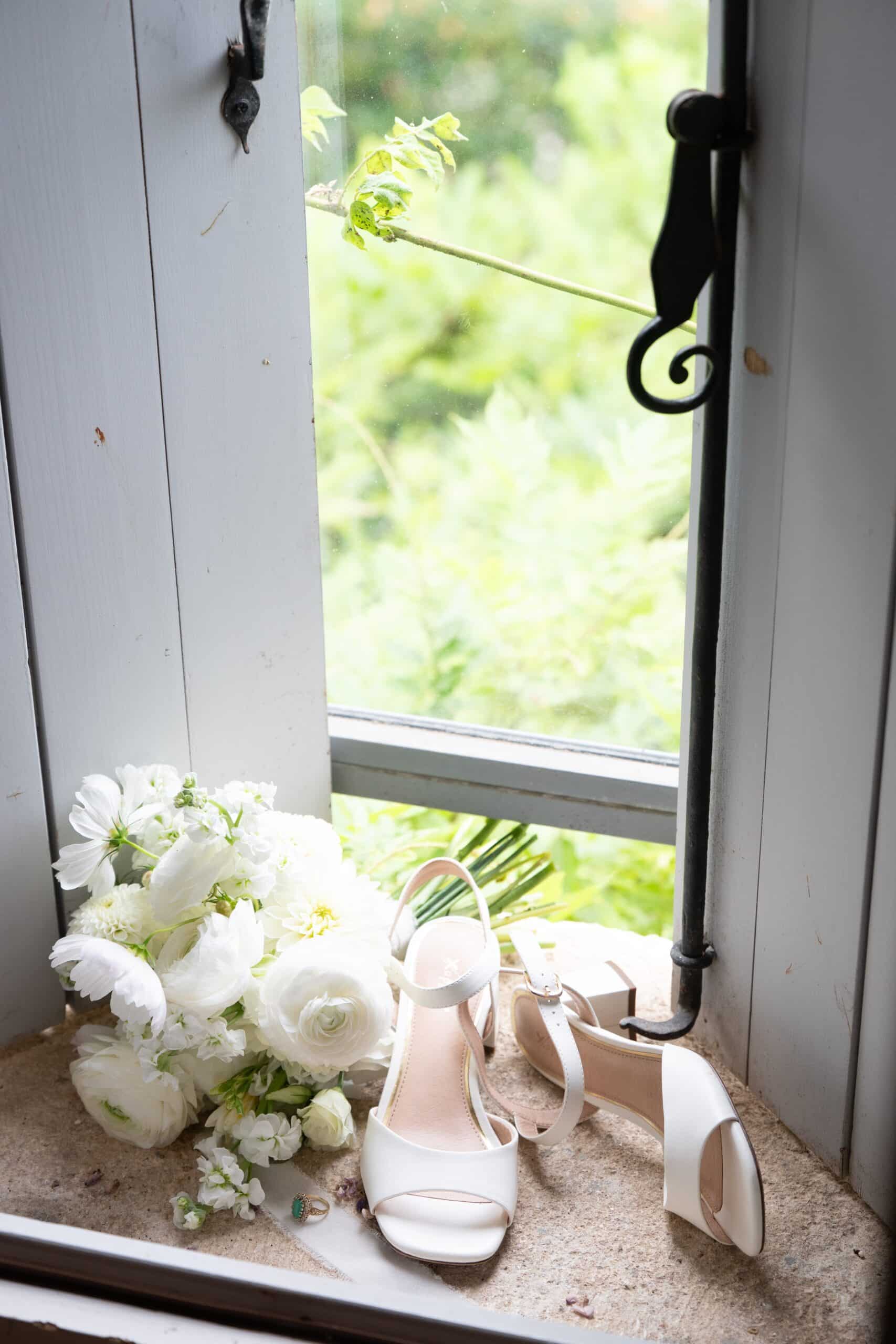 Bouquet de mariage blanc (ranunculus), chaussures de mariée blanches et bague en émeraude sur un rebord de fenêtre rustique.