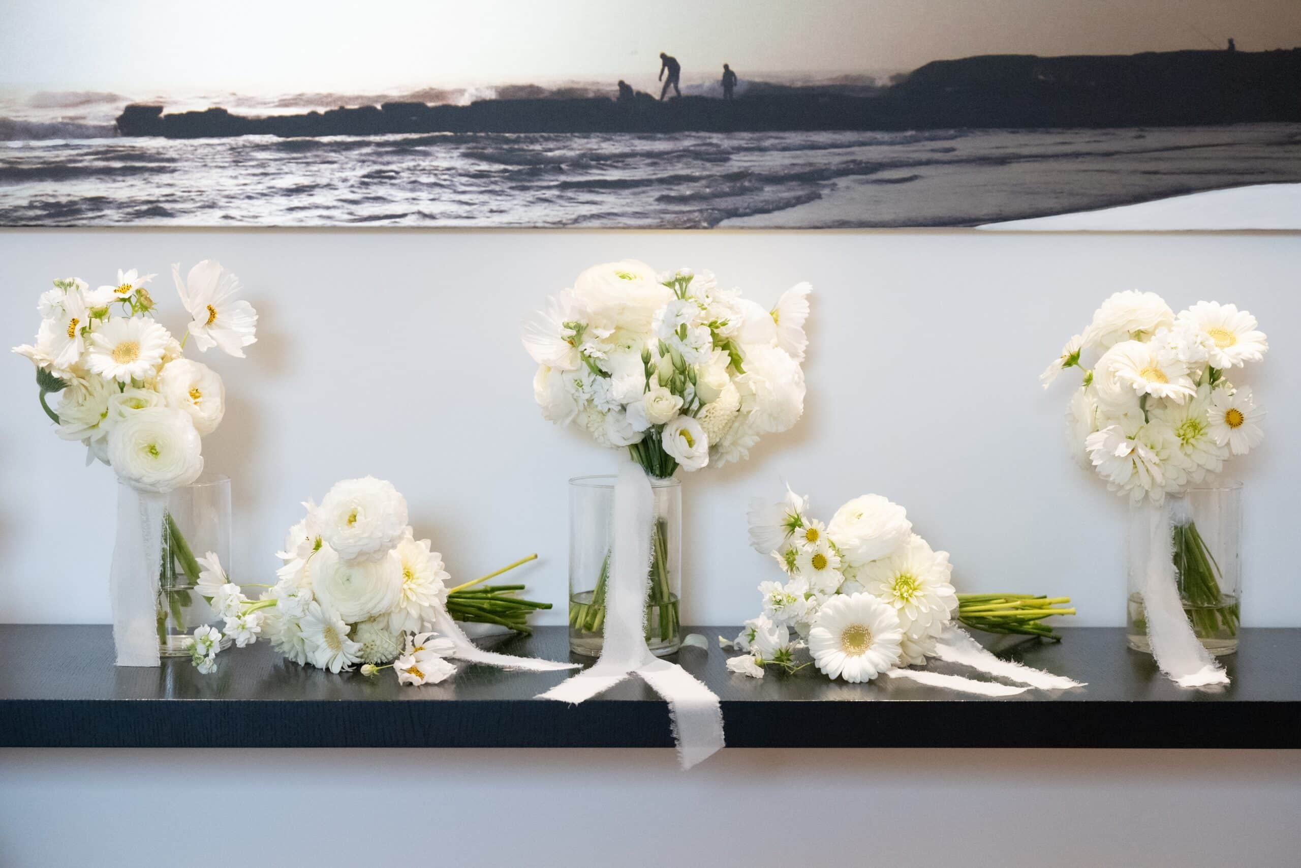 Arrangements de bouquets blancs (ranunculus, dahlias) sur étagère noire. Rubans et toile d'art panoramique d'océan et silhouettes.