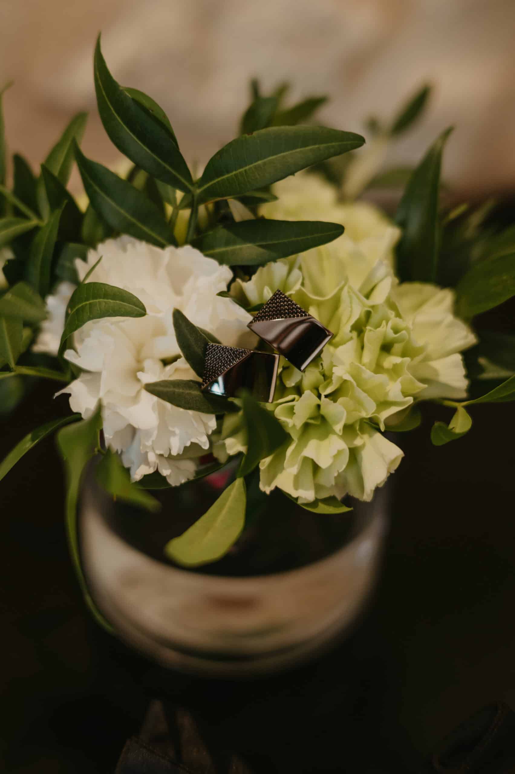 Boutons de manchette géométriques en métal noir texturé et chromé, posés sur un bouquet de mariage de fleurs blanches et vert clair.
