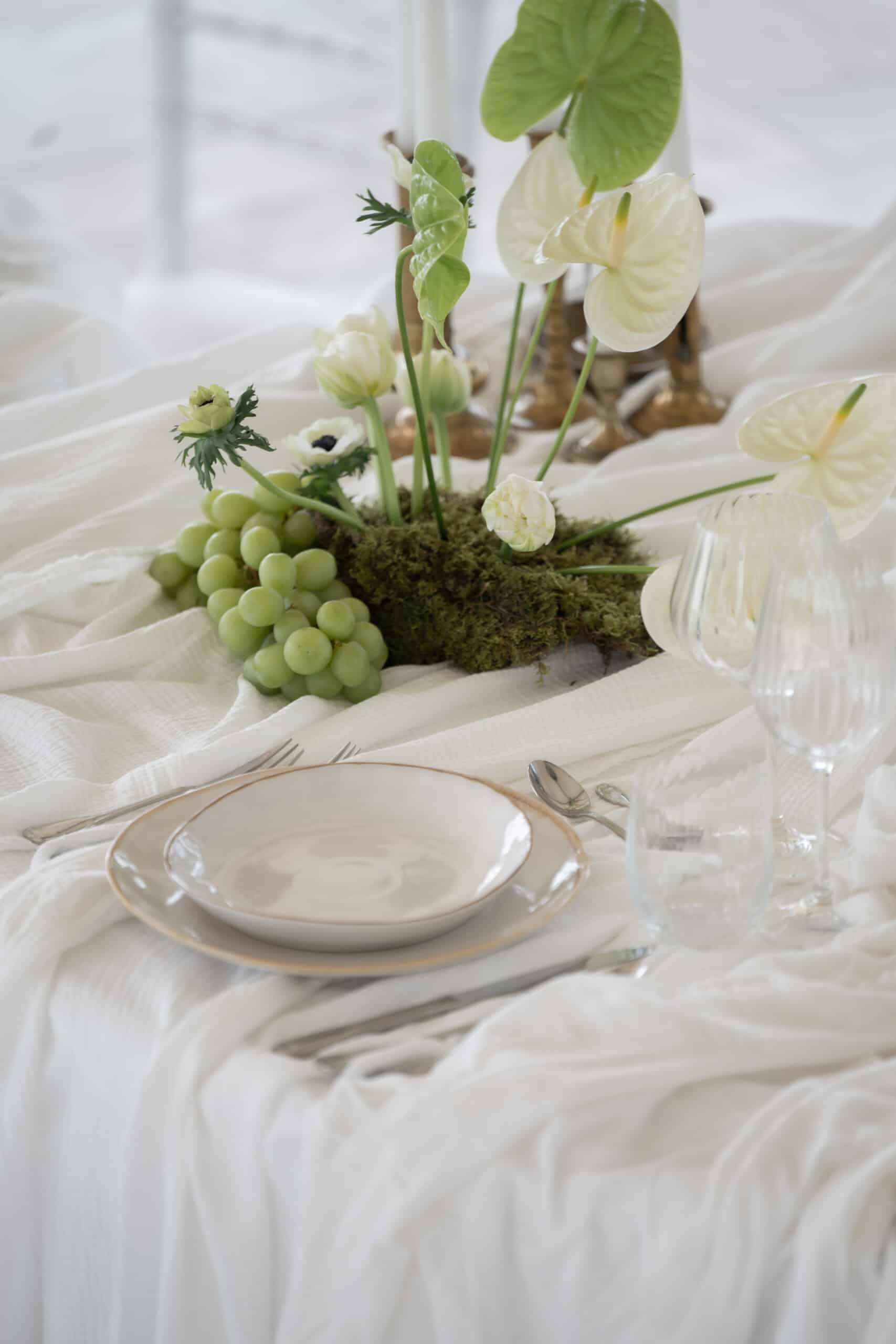 Décoration de table de mariage naturelle. Centre de table floral en mousse, anthuriums blancs, anémones et raisins verts sur nappe texturée.