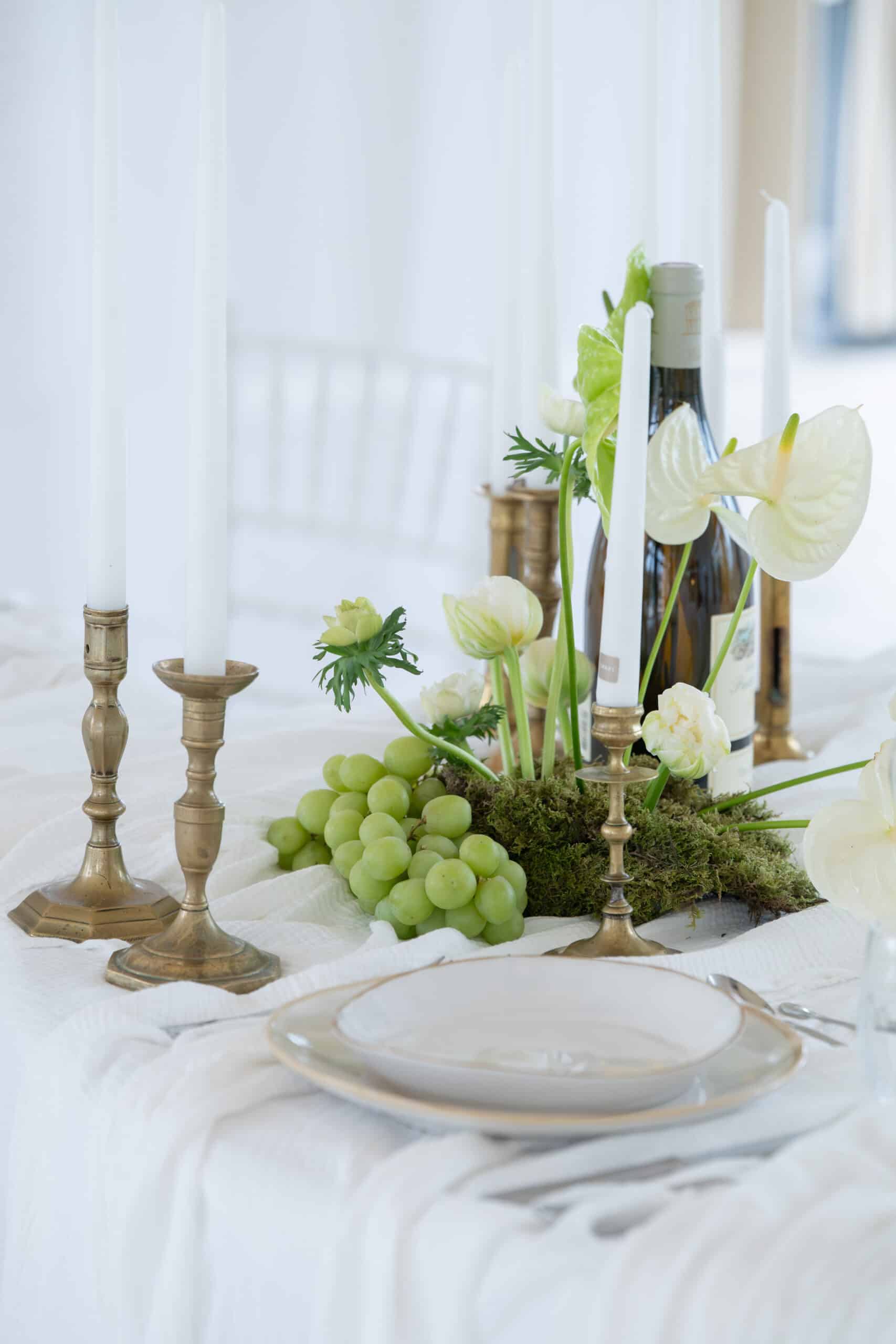 Centre de table élégant avec grappes de raisins verts, mousse, bougies blanches en chandeliers de laiton, fleurs blanches et bouteille de vin sur nappe texturée.