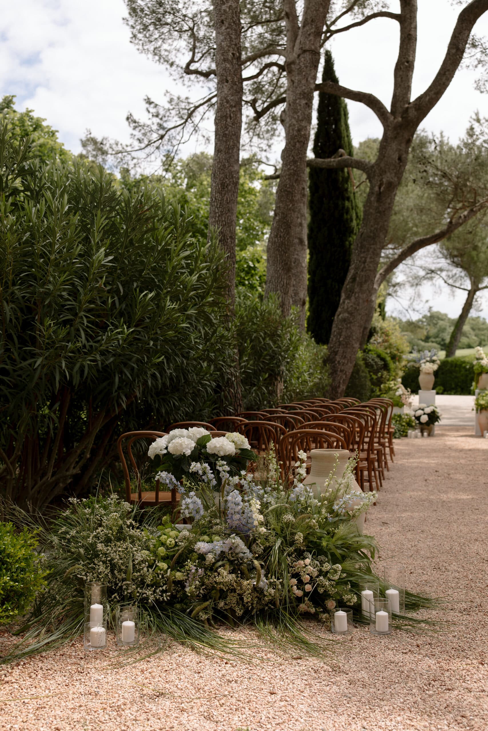 Allée de cérémonie de mariage extérieure bordée d'arbres. Rangées de chaises en bois, avec une riche décoration florale d'hydrangées et bougies.