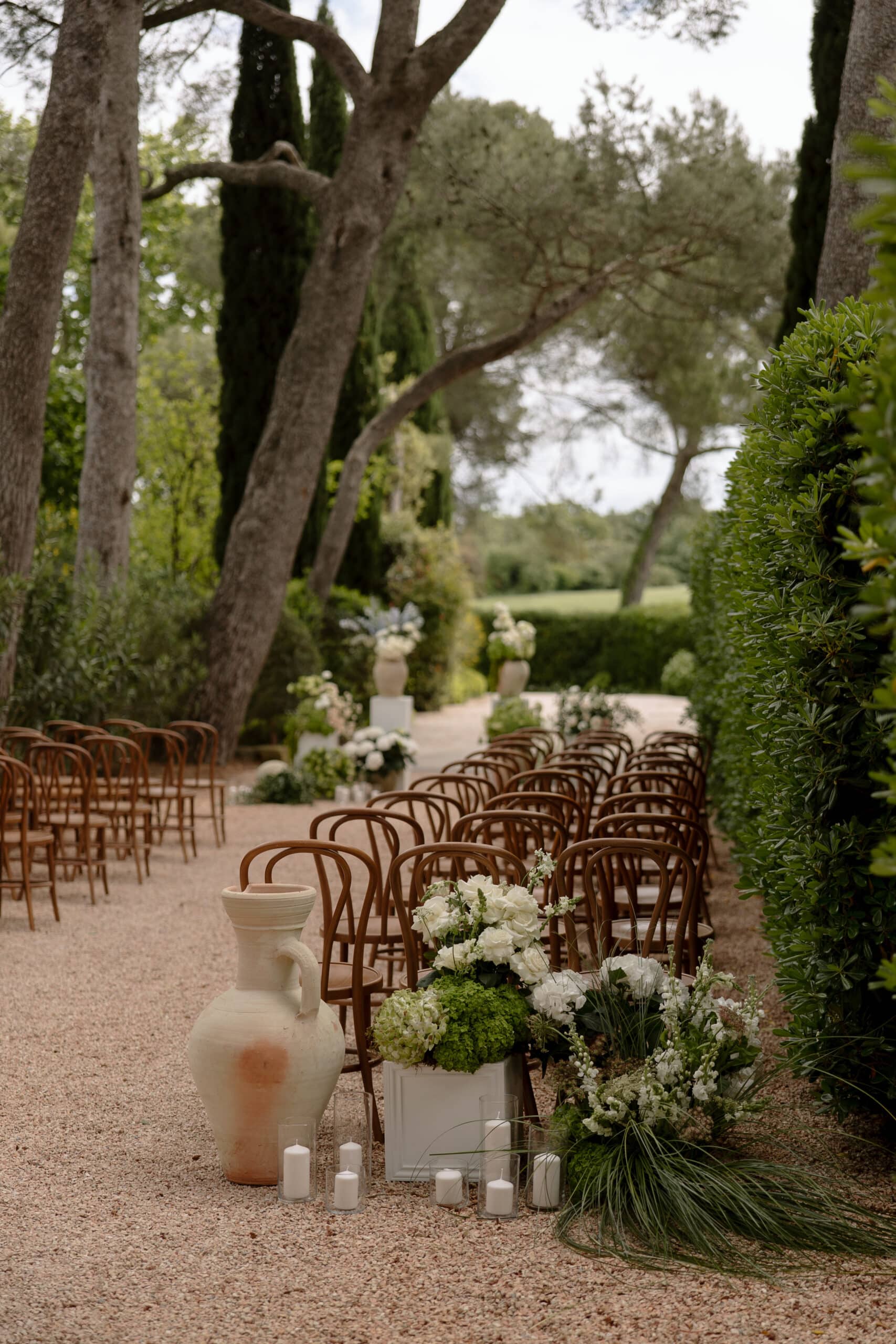 Allée de cérémonie de mariage extérieur dans un jardin. Chaises en bois courbé, vase rustique, bougies et arrangements floraux blancs élégants.