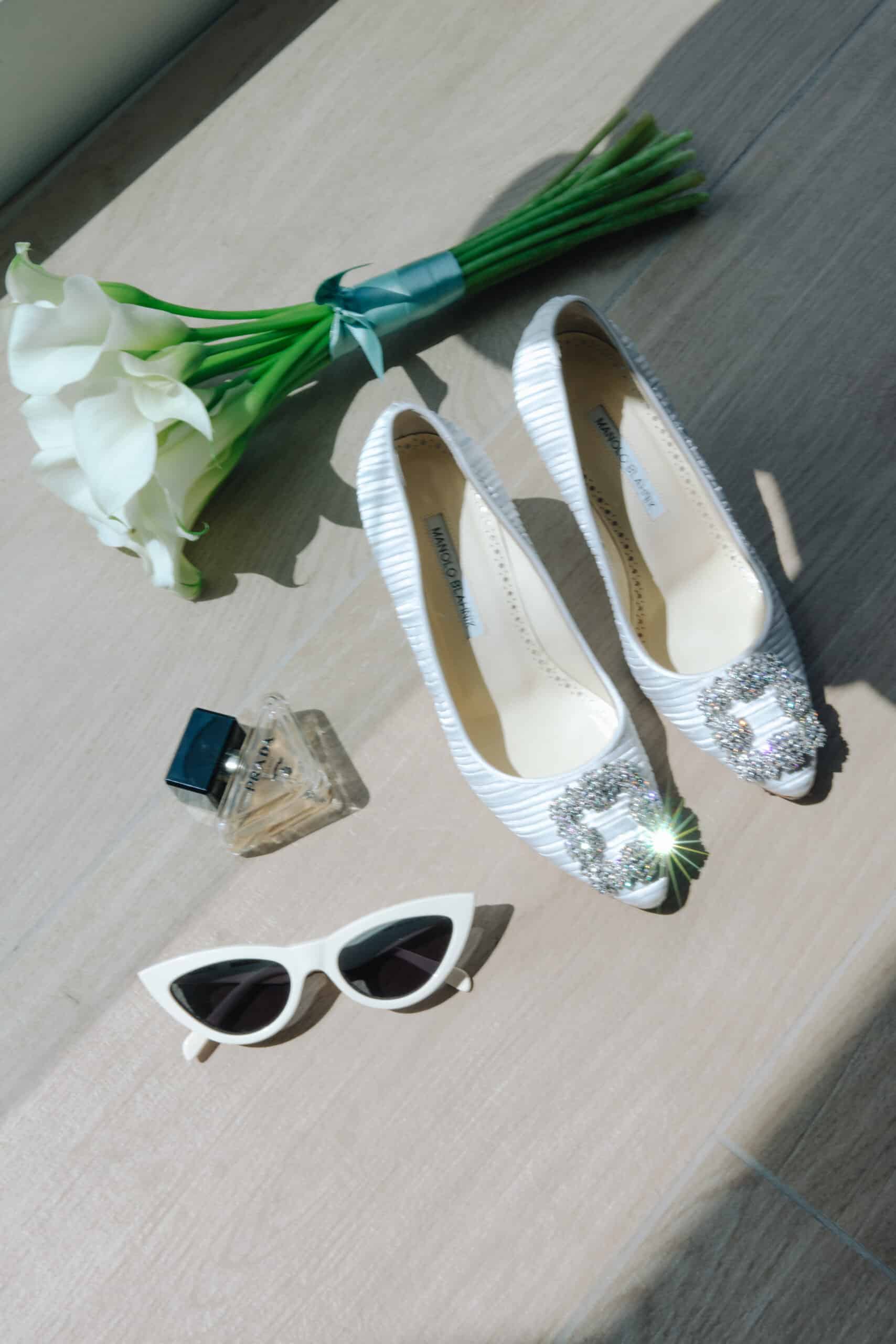 Chaussures de mariage Manolo Blahnik à boucle bijou, bouquet de callas blanches, parfum Prada et lunettes sous un soleil éclatant.