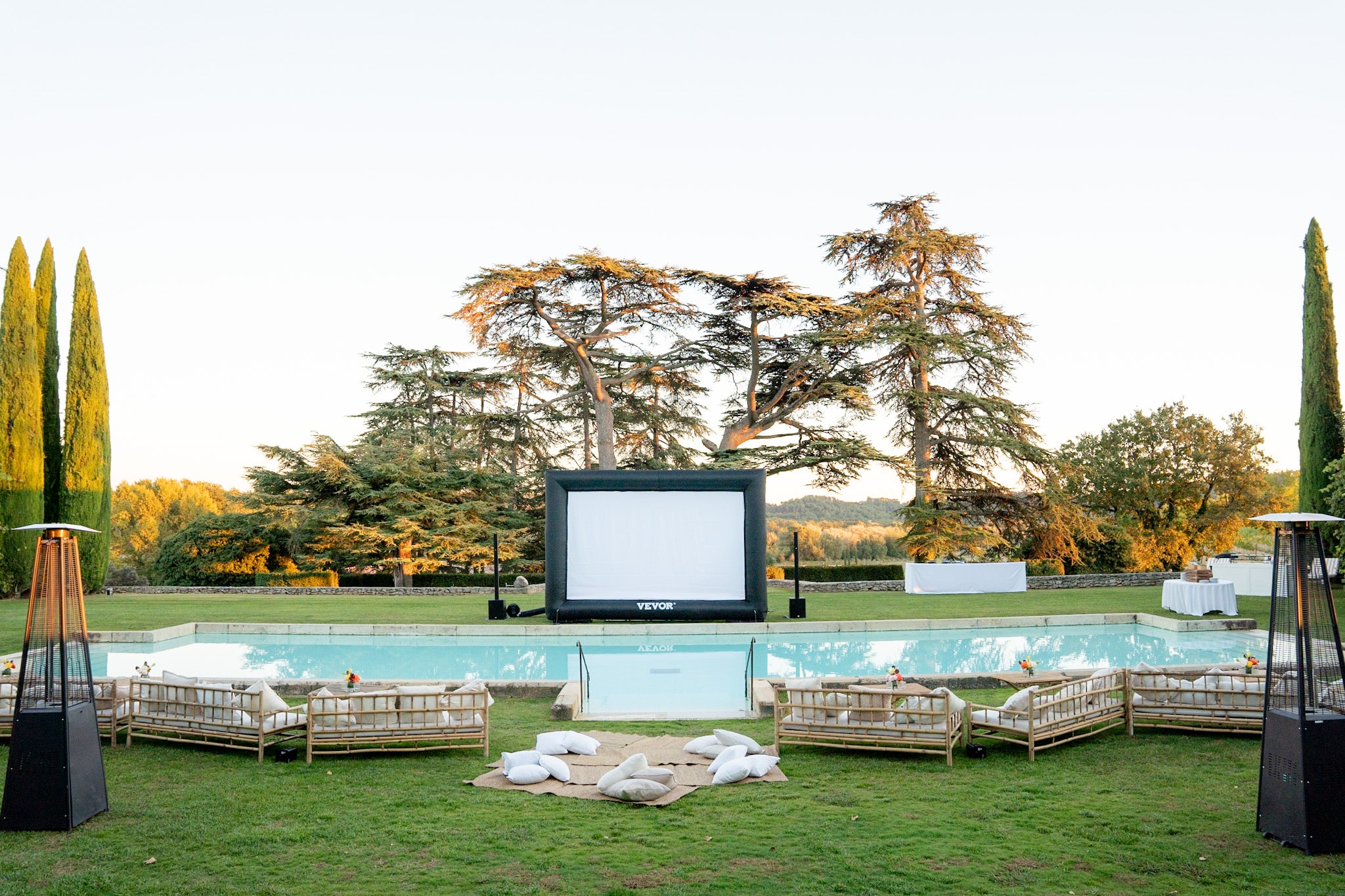 Cinéma extérieur de luxe au bord de la piscine. Écran VEVOR, canapés en bambou et chauffe-terrasses sous la lumière dorée du soir.