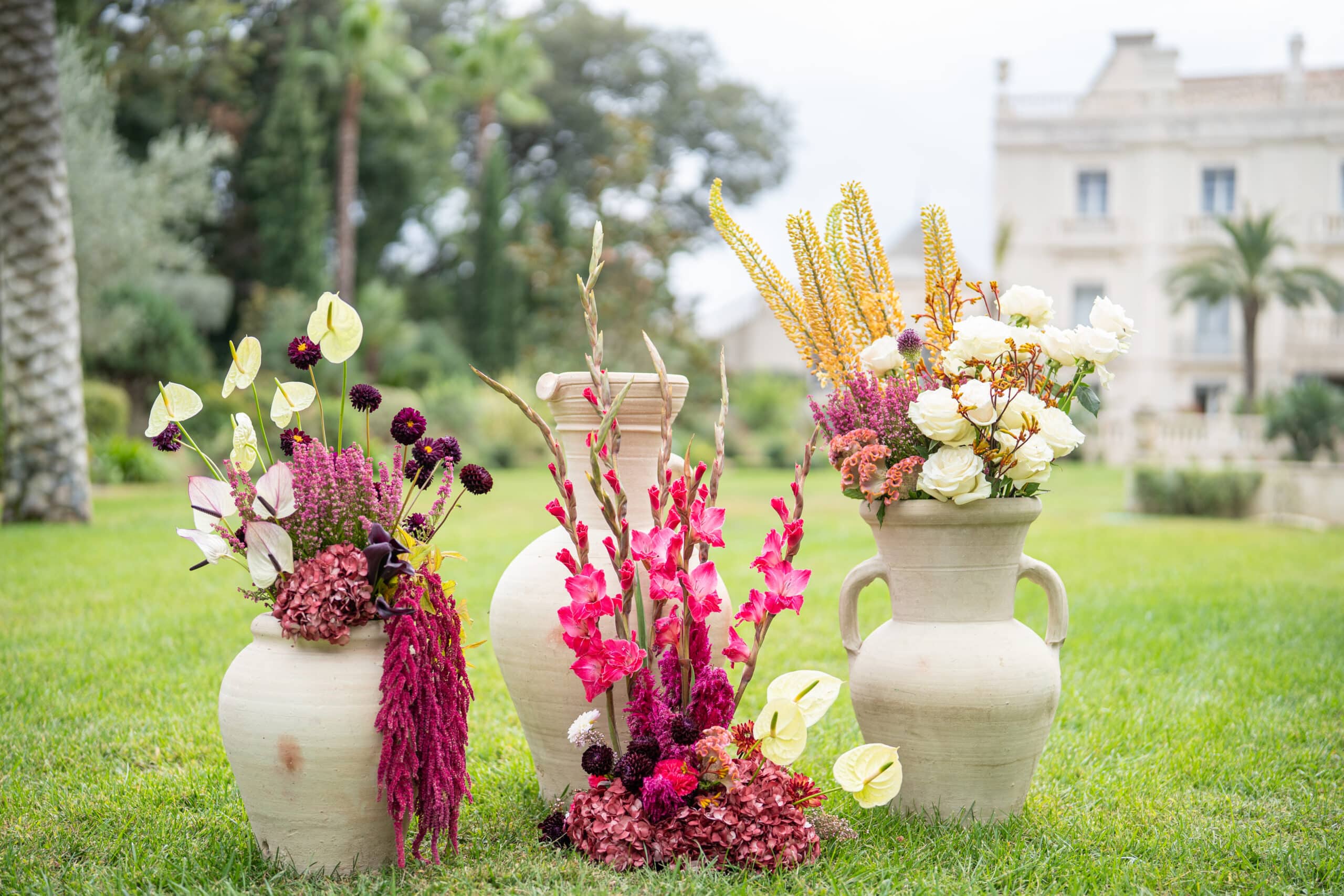 Vases en terre cuite sur gazon, ornés de compositions florales magenta, pourpres et blanches (roses, anthuriums) pour une décoration de mariage luxe en extérieur.