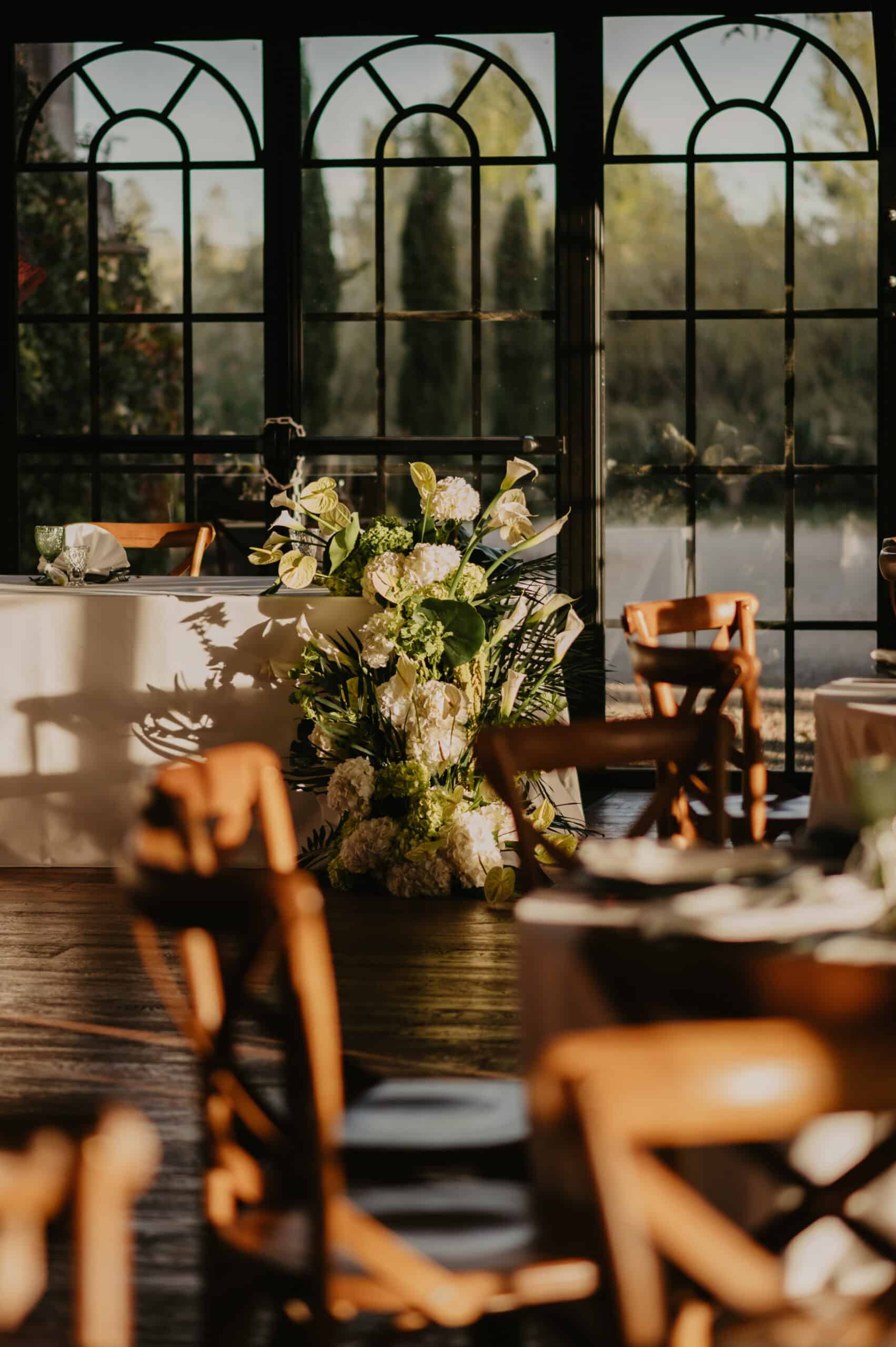 Scène de réception de mariage élégante. Fenêtre à arceaux baignée de lumière, projetant des ombres. Centrée sur un bouquet d'arums et d'hydrangées blancs.
