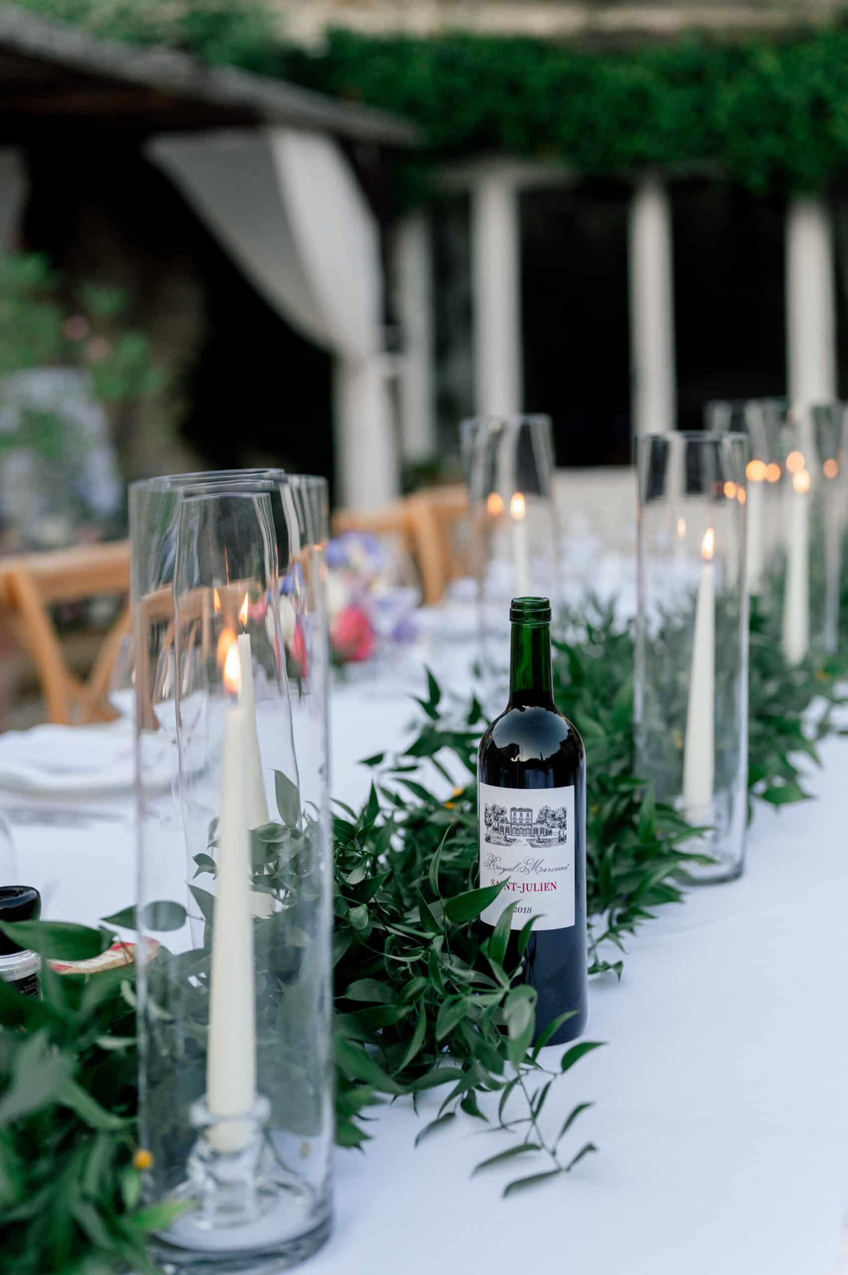 Longue table de réception avec guirlande de verdure, bougies allumées sous verre et vin Saint-Julien Royal Marceau 2018, créant une ambiance élégante.