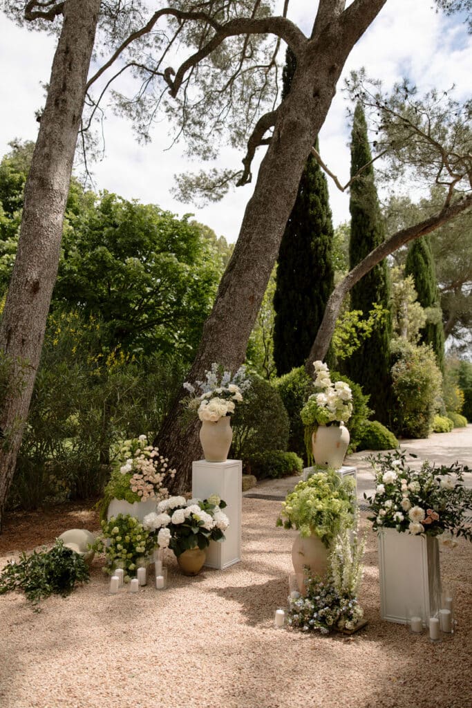 Magnifique décor floral de cérémonie de mariage en extérieur. Vastes arrangements de roses blanches et de verdure dans des vases rustiques, sous de grands arbres.