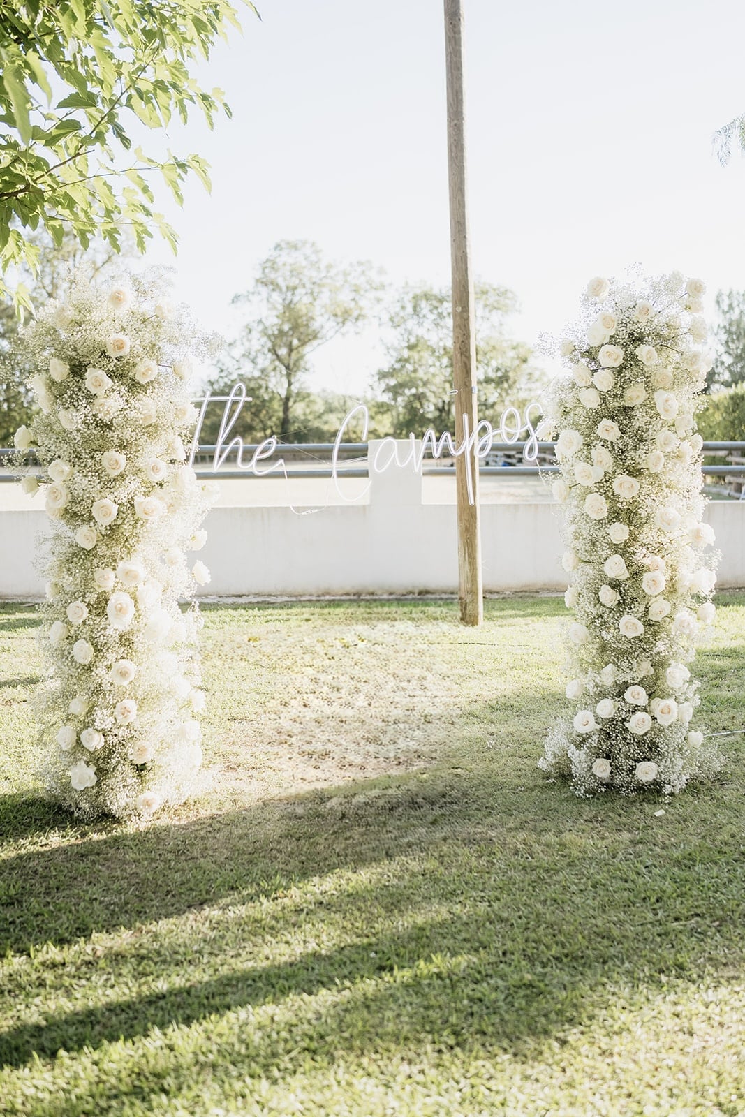 Deux colonnes fleuries de roses et gypsophile créent une arche extérieure. Enseigne néon "The Campos" illuminée.