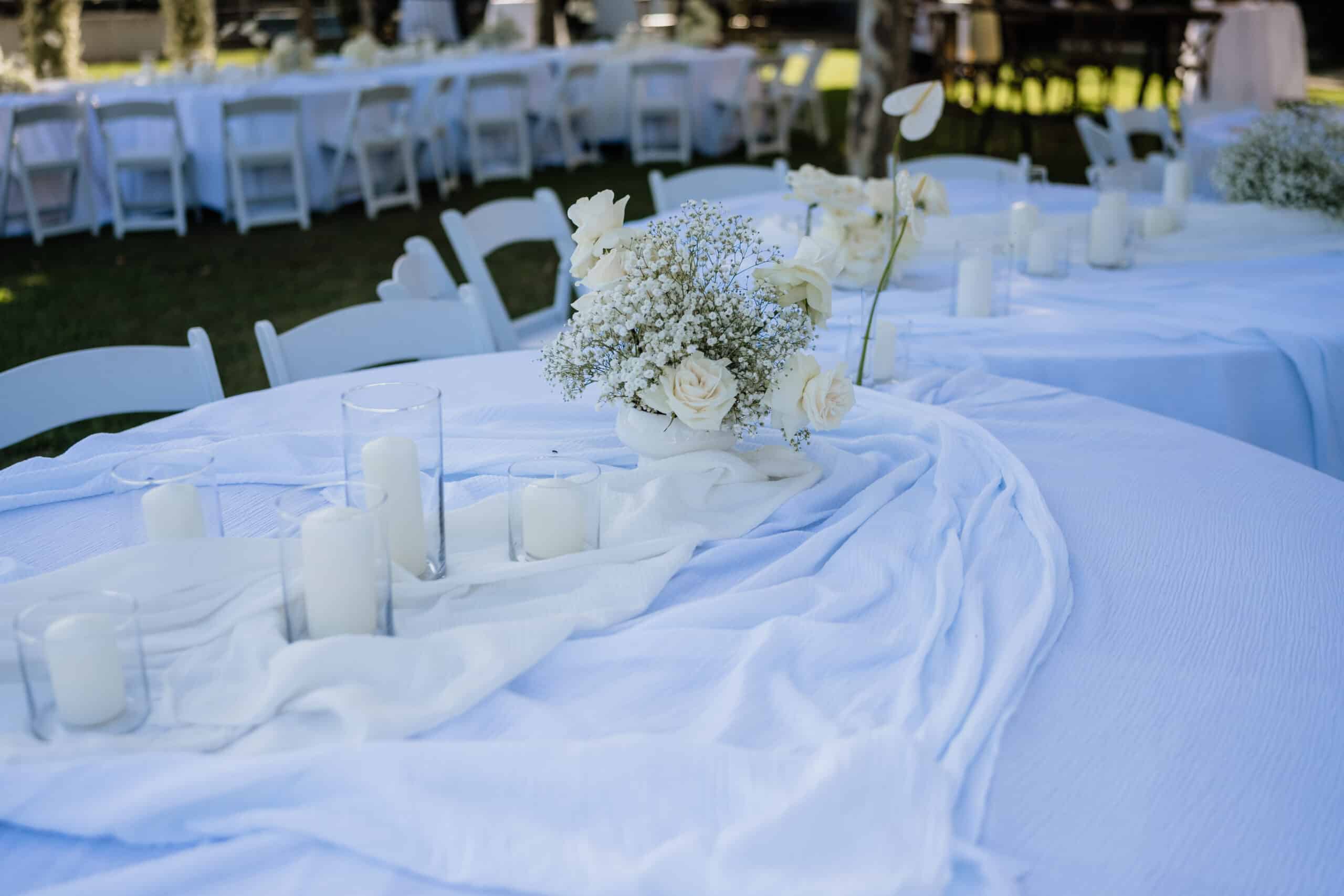 Décor de table de mariage extérieur : nappe bleue, chemin de table, roses blanches, gypsophile et bougies. Chaises blanches en arrière-plan.