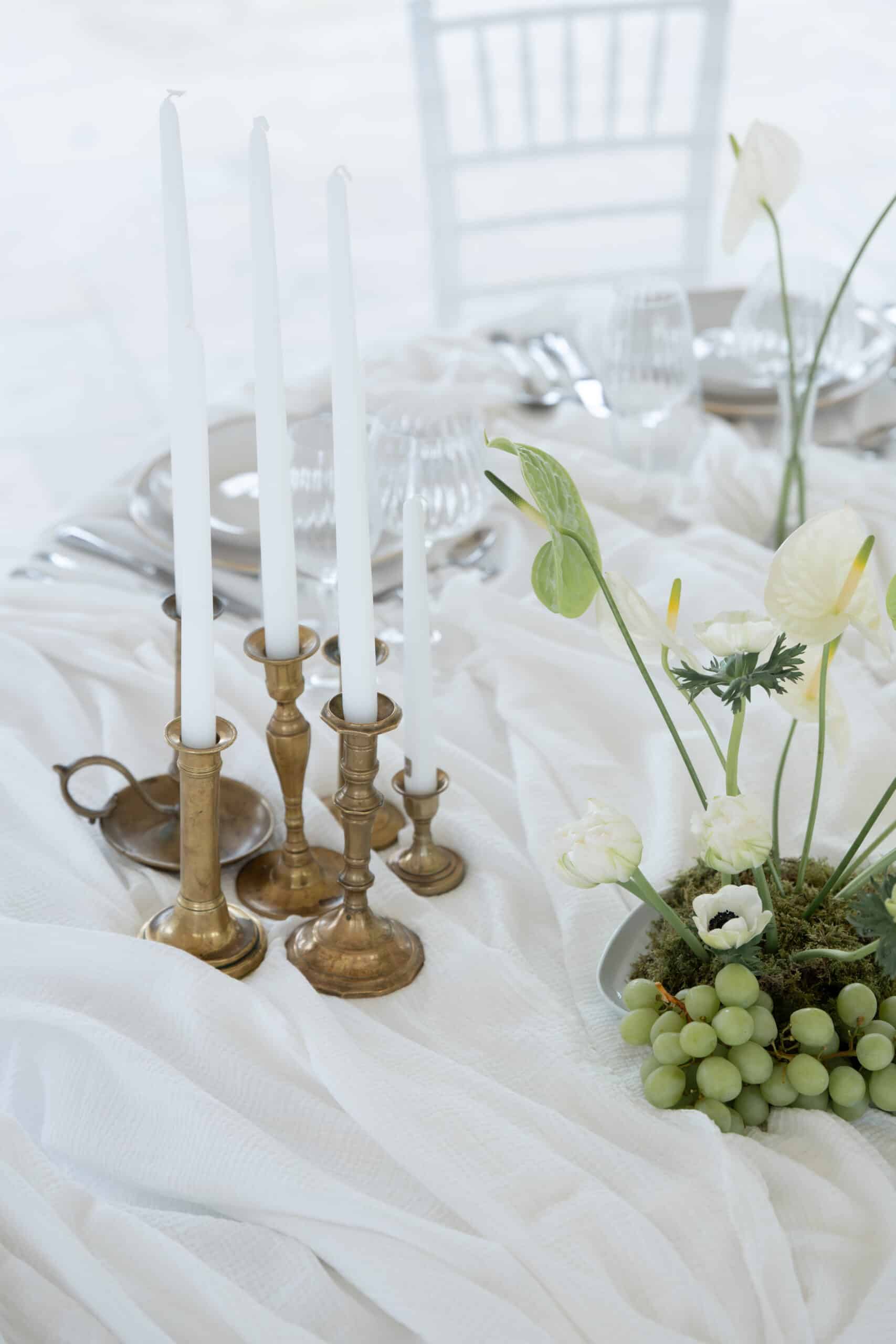 Table de fête monochrome : bougies blanches dans des chandeliers en laiton, centre de table floral (anthuriums) et raisins sur nappe blanche élégante.