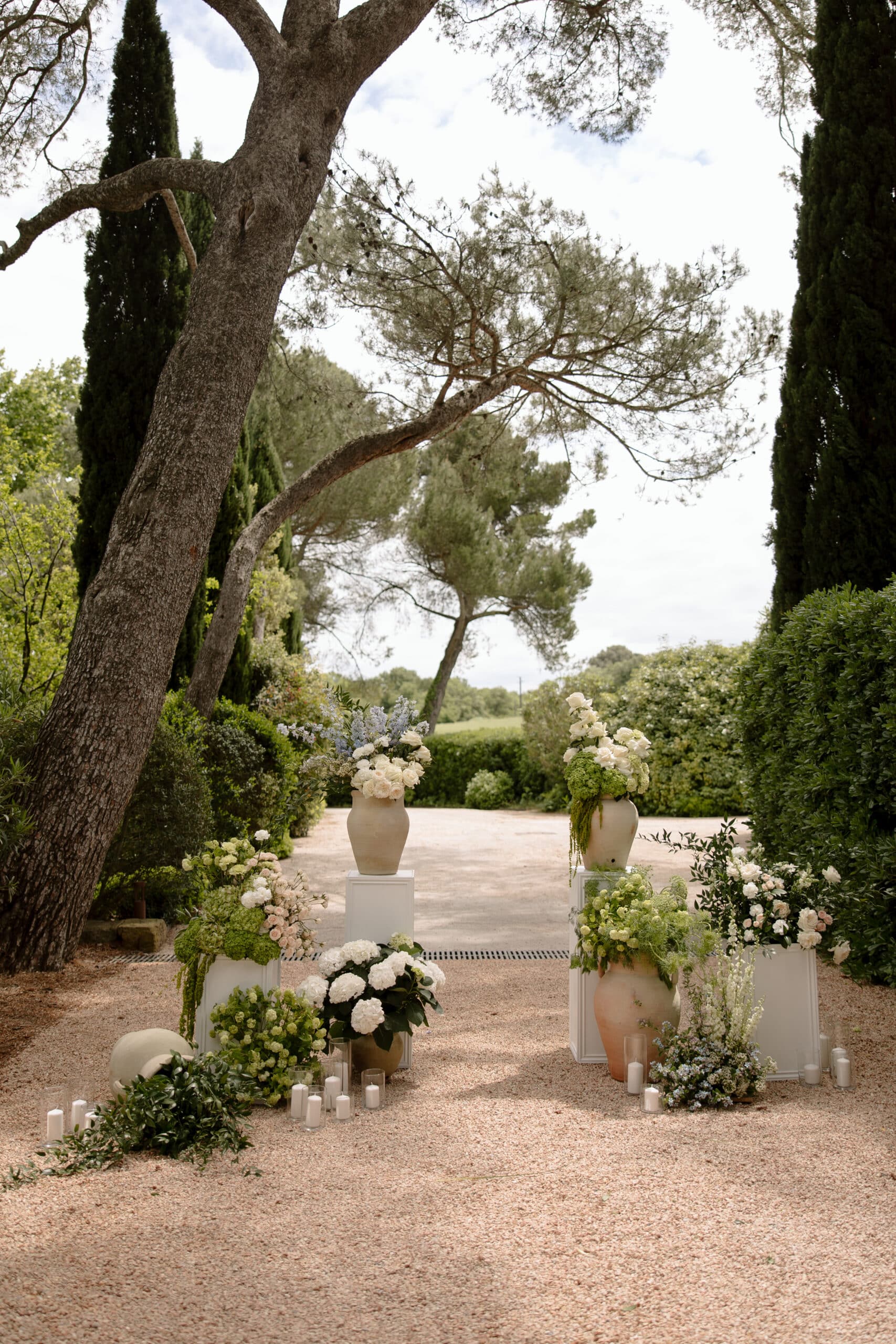 Élégante décoration florale de mariage encadrant une allée de jardin. Vases en céramique, compositions blanches et vertes, et bougies sur gravier.