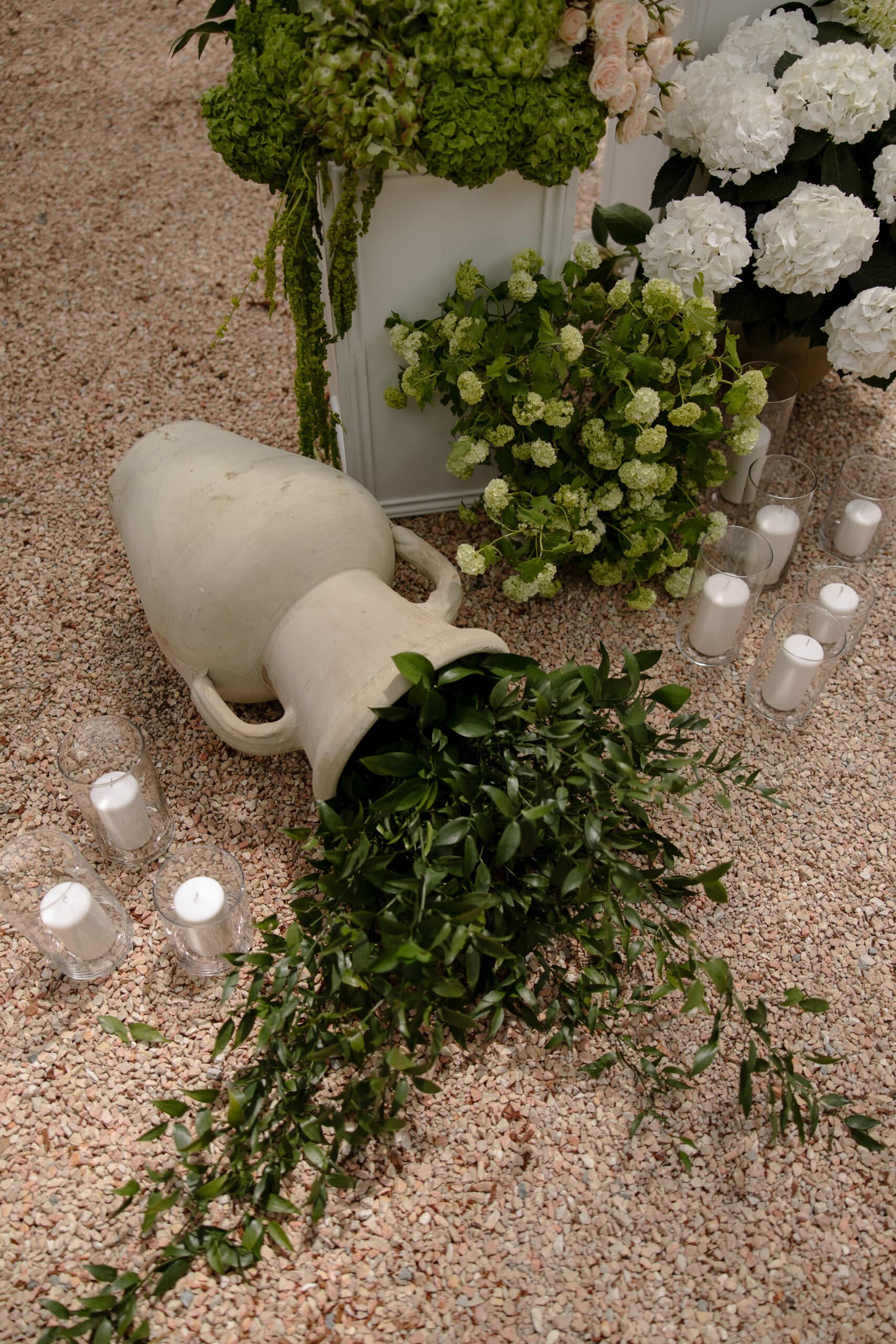 Amphore crème renversée sur gravier, verdure tombante. Décoration de mariage extérieure avec bougies cylindriques, hortensias blancs et verts.