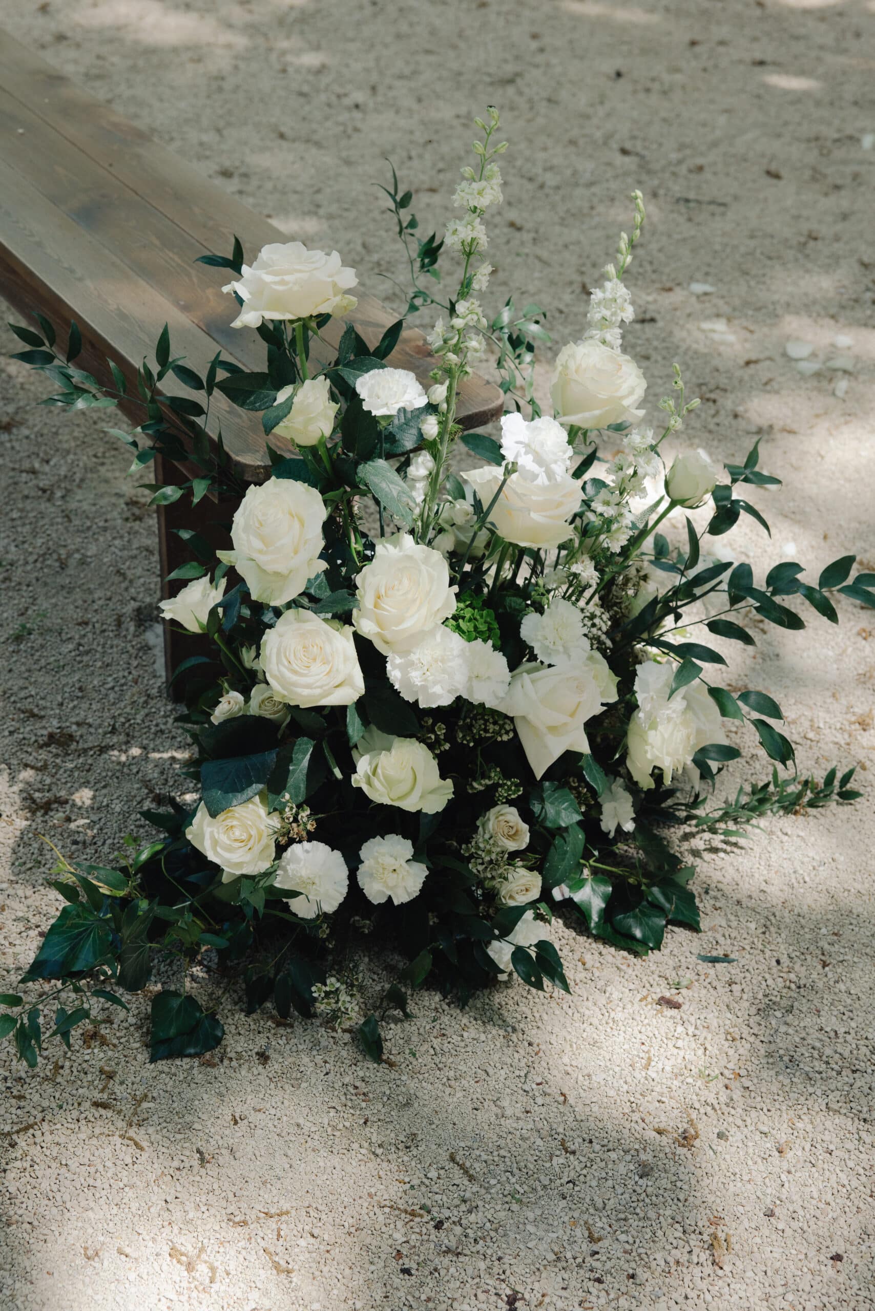 Magnifique décoration de mariage : roses blanches, œillets et feuillage vert sur un banc en bois rustique, posée sur du gravier.