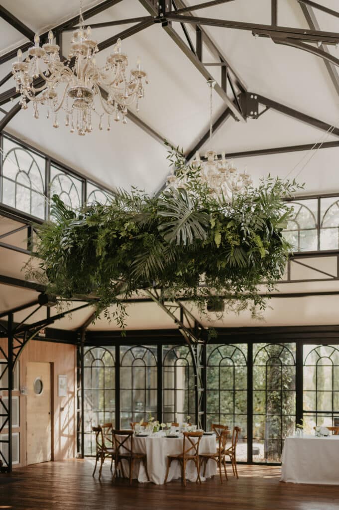 Réception de mariage sous une verrière industrielle. Les tables sont décorées sous une suspension de verdure luxuriante et deux lustres en cristal.