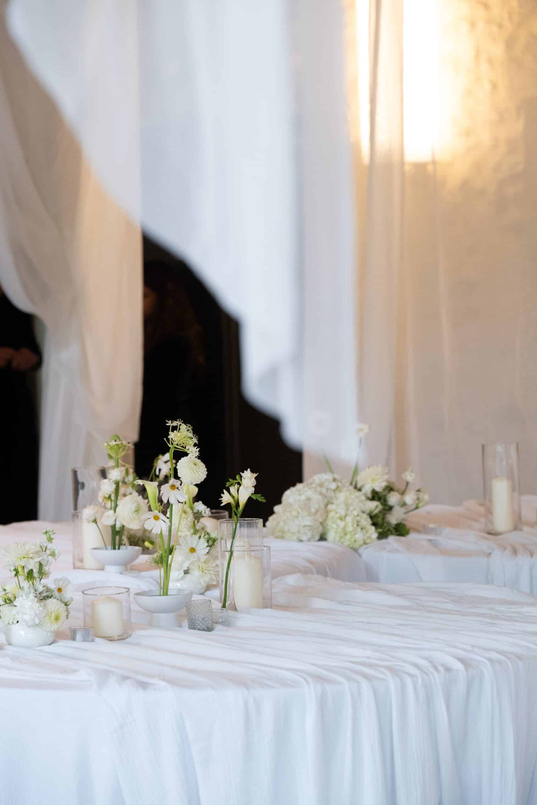 Décoration élégante de mariage. Longue table drapée de tissu blanc, arrangements de fleurs blanches et bougies. Ambiance douce et lumineuse.