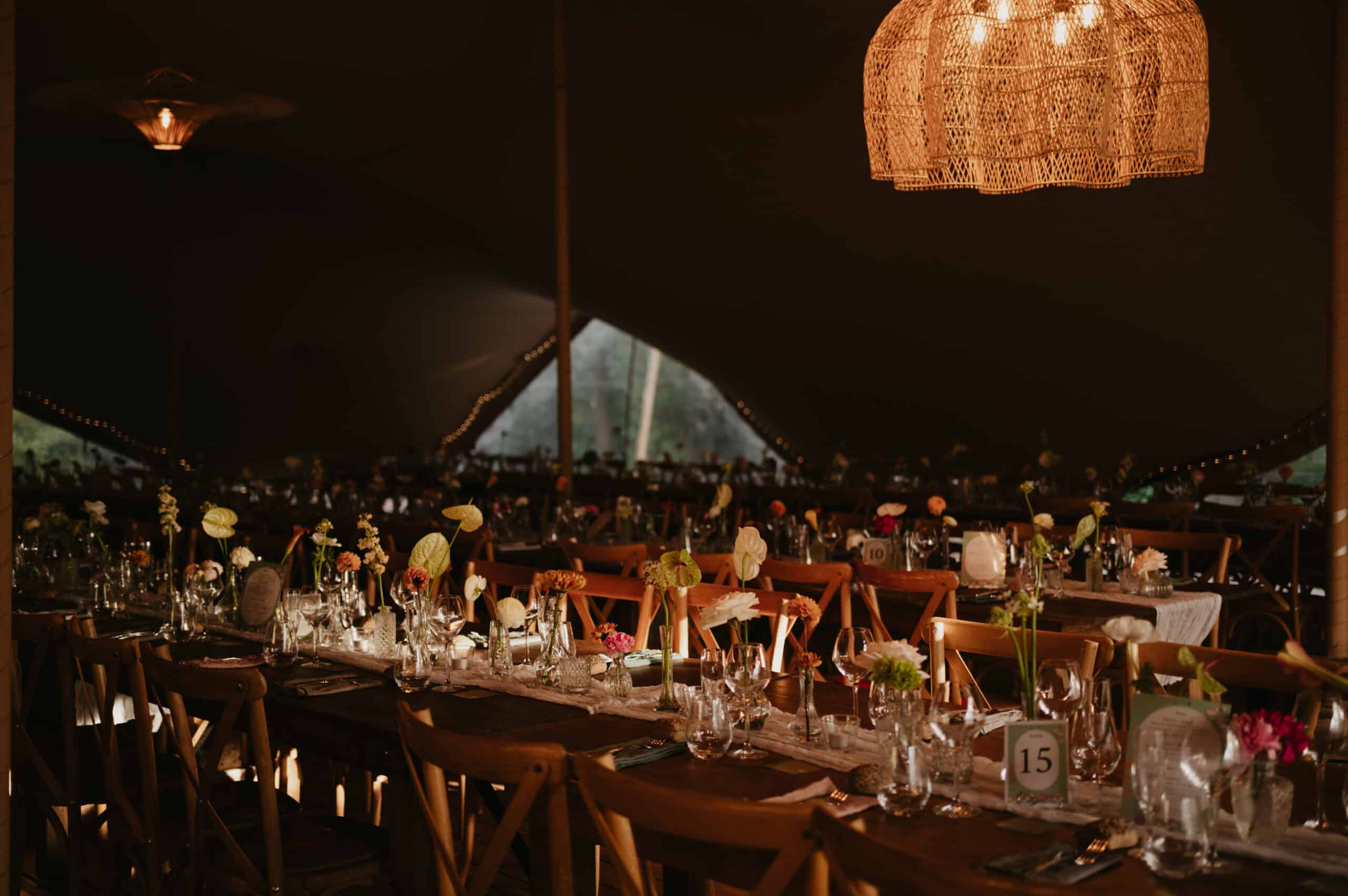 Atmosphère chaleureuse sous tente de mariage. Tables rustiques élégantes dressées avec verrerie, décorations florales et lumière dorée.