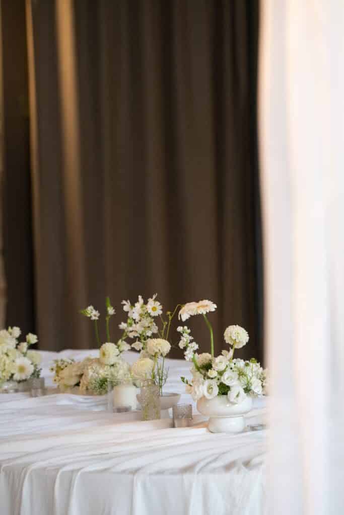 Centre de table élégant. Compositions de fleurs blanches variées et bougies sur nappe plissée devant de lourds rideaux marron foncé.
