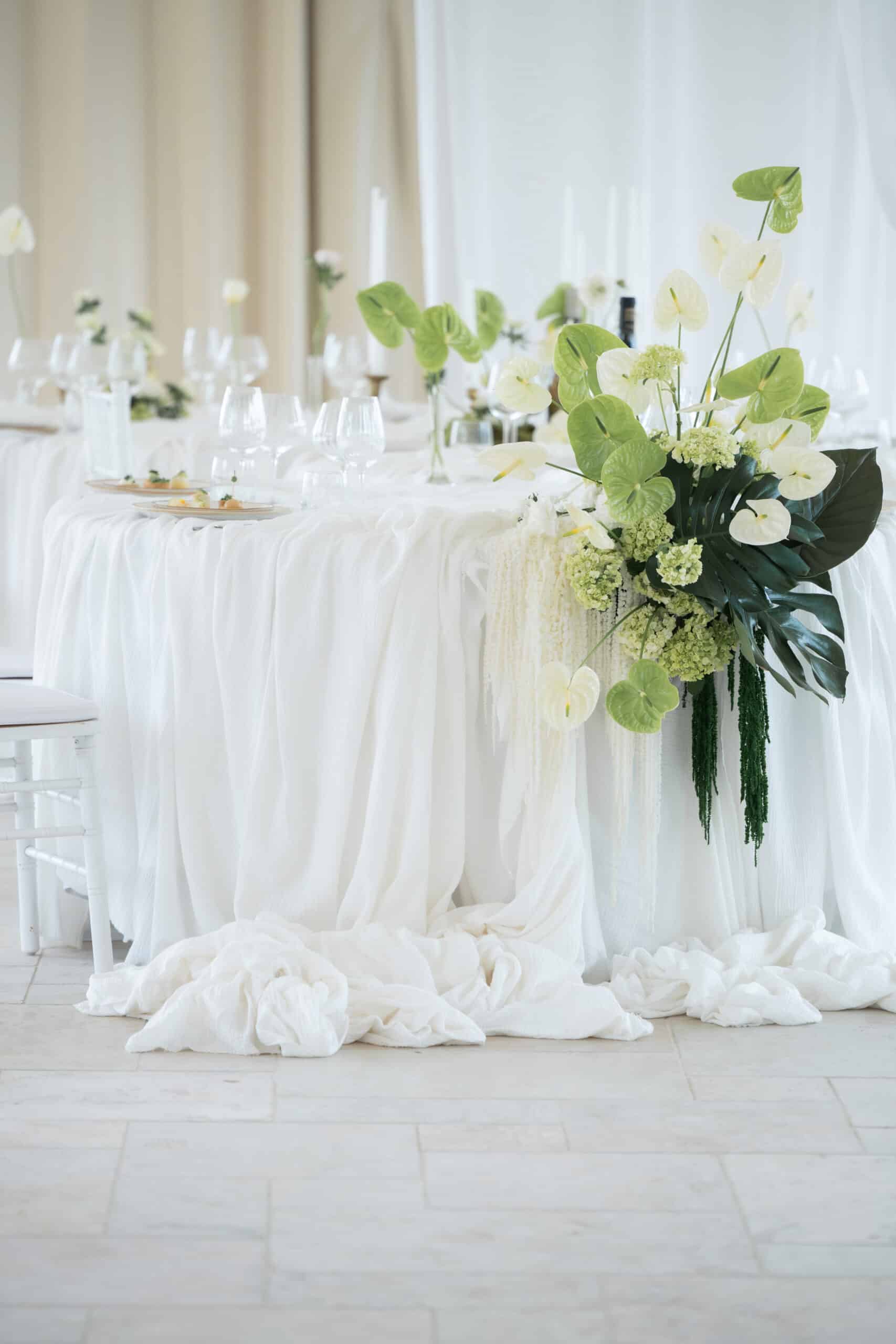 Décoration de table élégante: nappe blanche fluide, Anthuriums verts et blancs, Monstera, et bougies pour une réception chic.