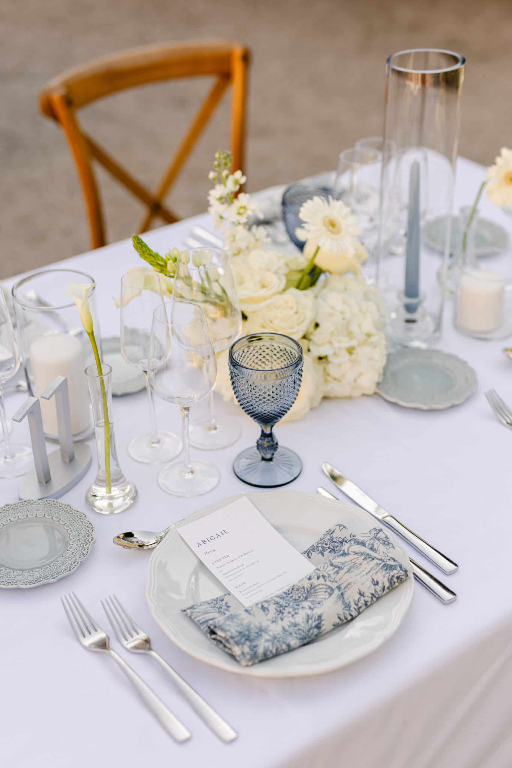 Table de mariage extérieure: centre de roses blanches, goblet bleu, bougies grises. Menu "ABIGAIL", serviette toile de Jouy et couvert argenté.