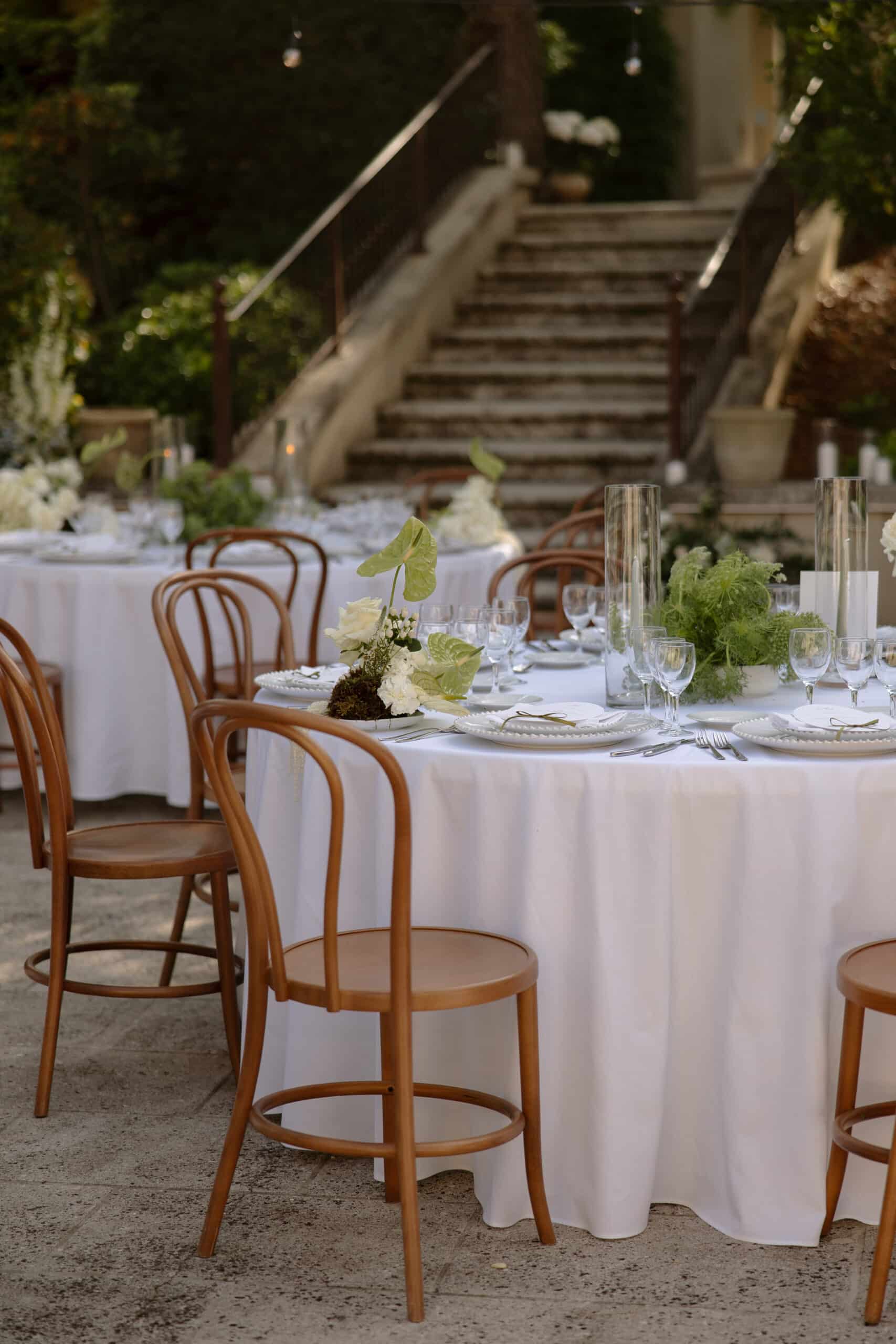 Table d'événement élégante en extérieur : nappe blanche, chaises en bois, centre de table roses blanches et anthurium vert, avec escalier en pierre en arrière-plan.