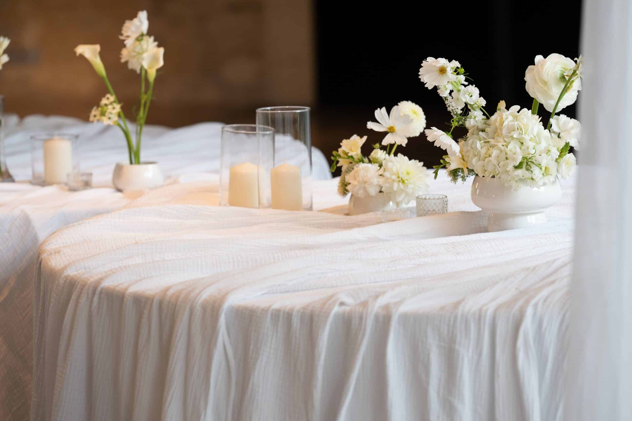 Table de mariage élégante couverte de lin blanc froissé. Décoration florale monochrome (lys, hydrangées, renoncules) et bougies.