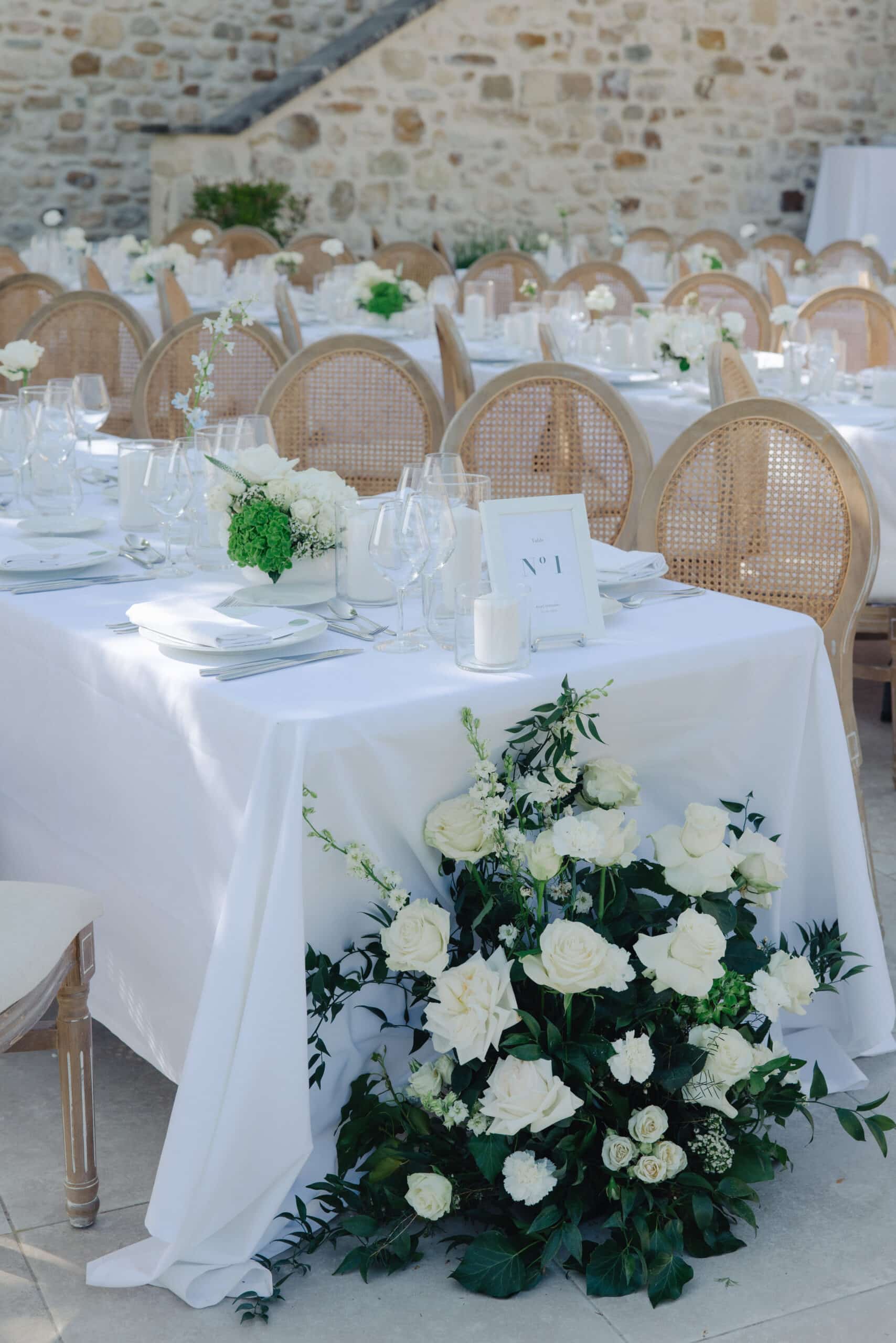 Réception de mariage extérieure chic. Table dressée de roses blanches, bougies, chaises en rotin. Table N° 1 pour Anna et Christopher.