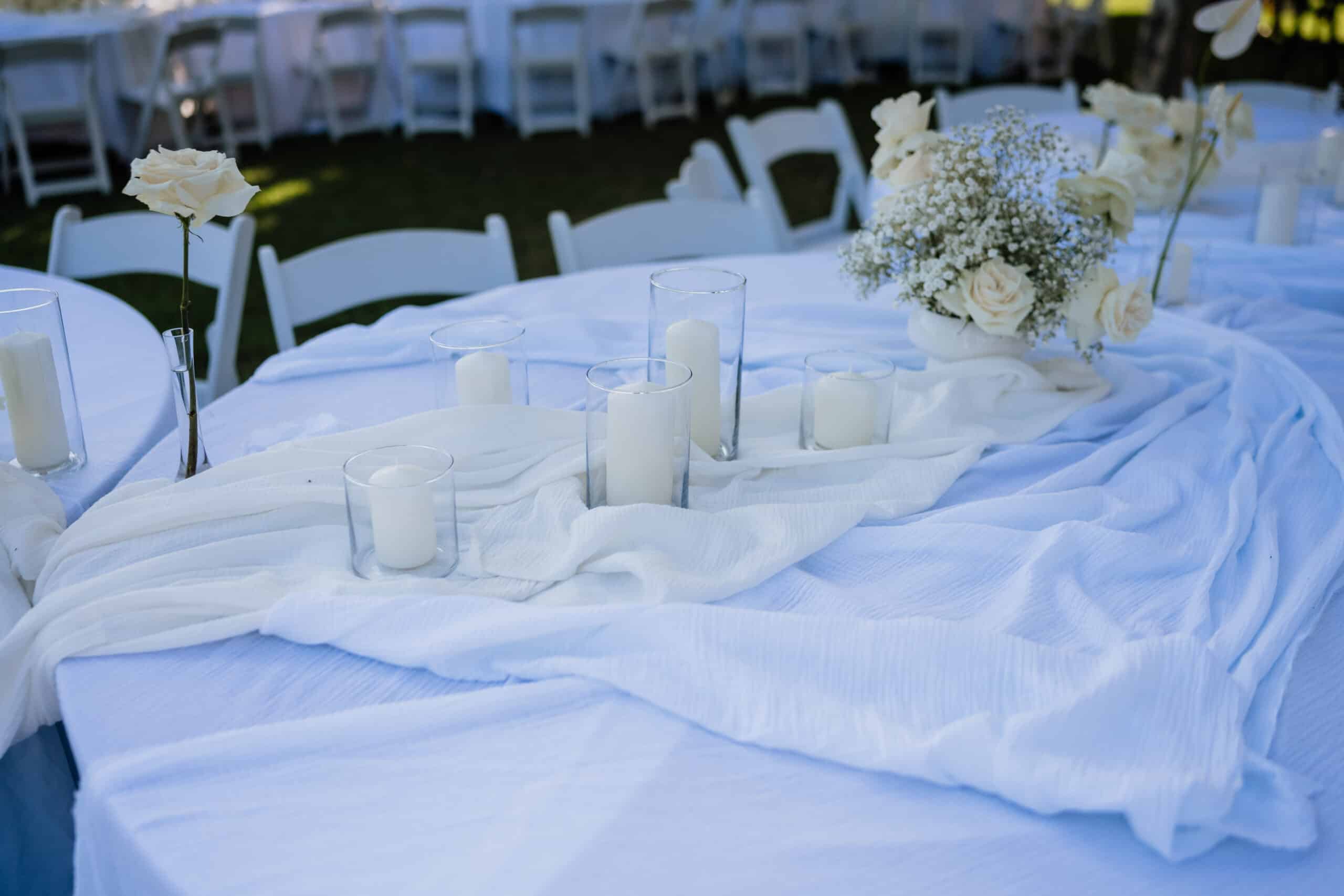 Centre de table de mariage extérieur : tissu drapé blanc, roses, gypsophile et bougies pilier dans des cylindres en verre transparent.