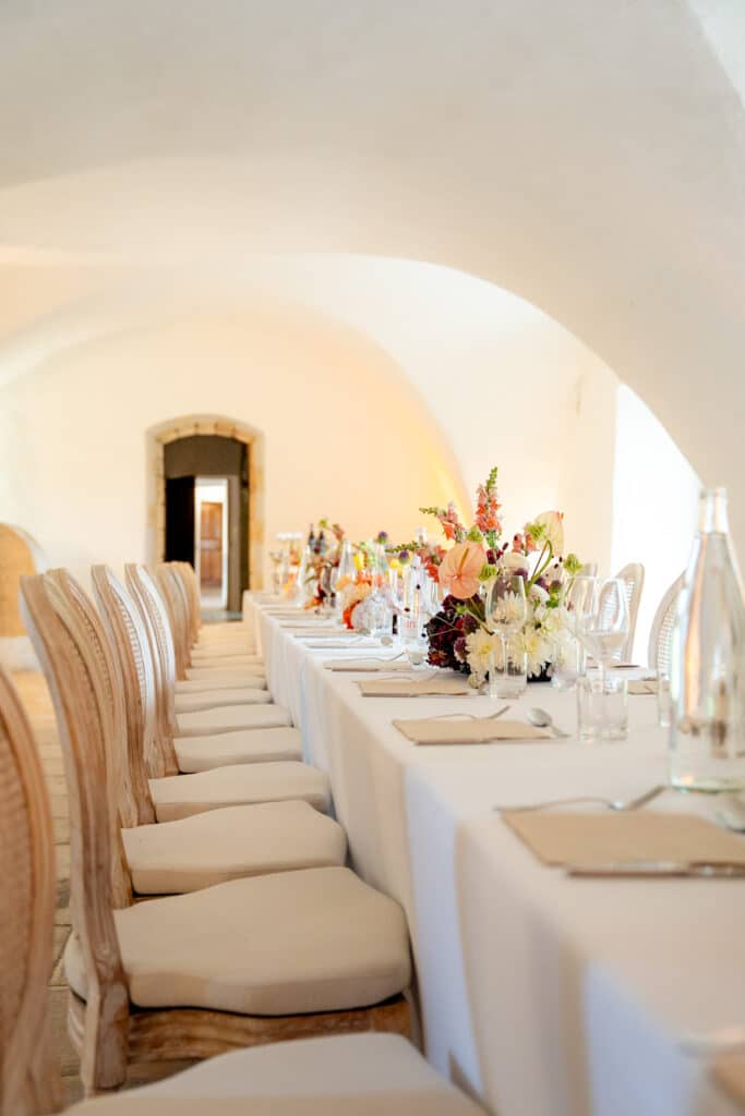 Longue table de réception de mariage dans une salle voûtée lumineuse. Chaises chics en bois, nappe blanche et centres de table floraux éclatants.