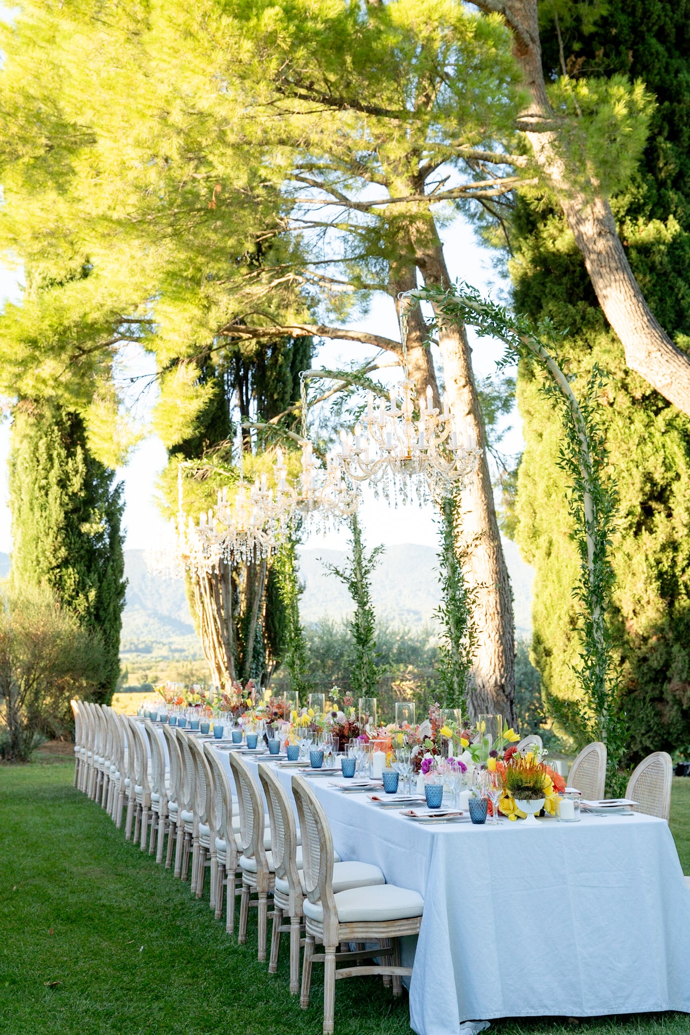 Longue table de réception de mariage élégante sous les arbres, décorée de lustres en cristal suspendus et de fleurs vibrantes.