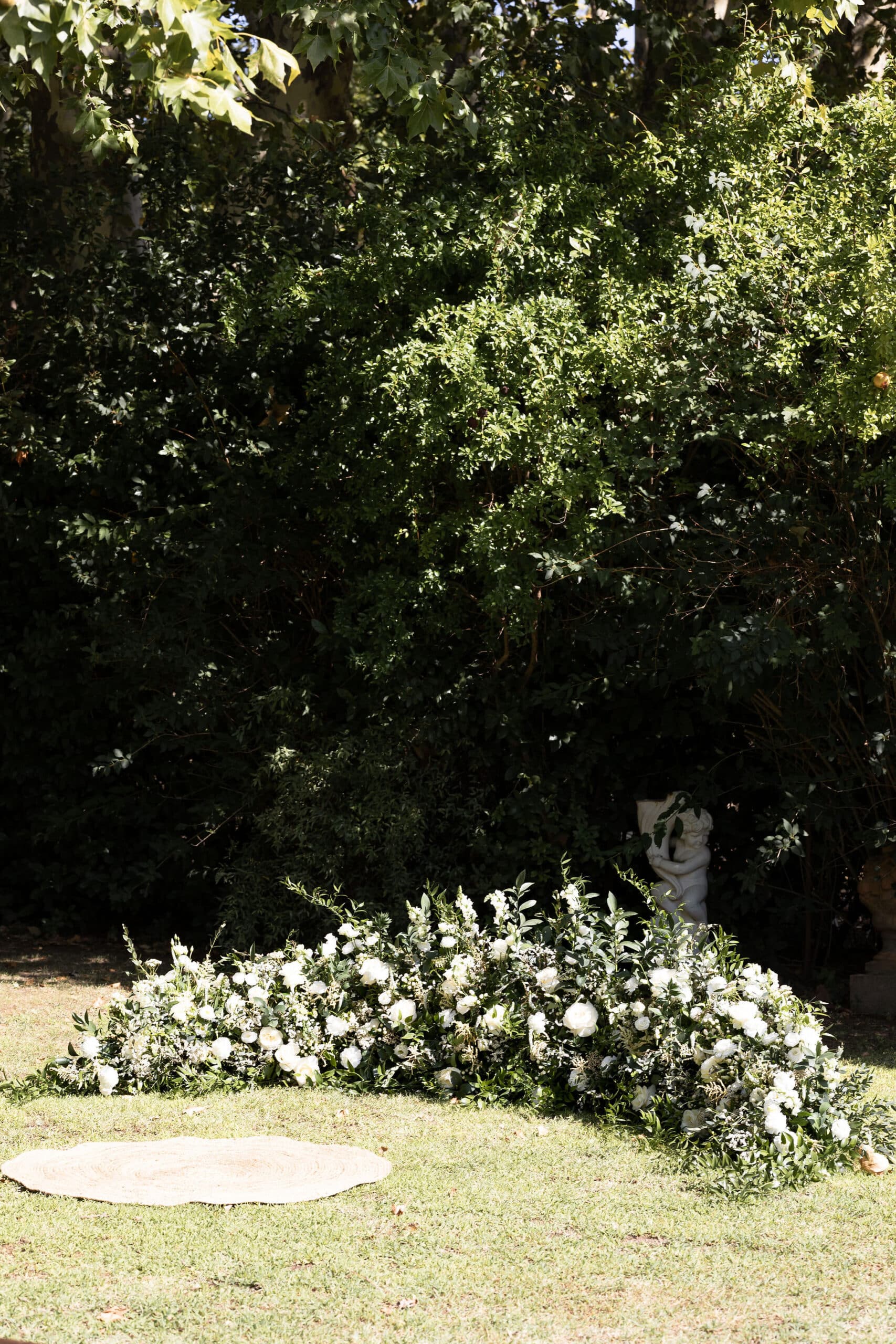 Arrangement bas de roses et fleurs blanches pour une cérémonie de mariage en jardin, avec un tapis en jute sur l'herbe et un chérubin.