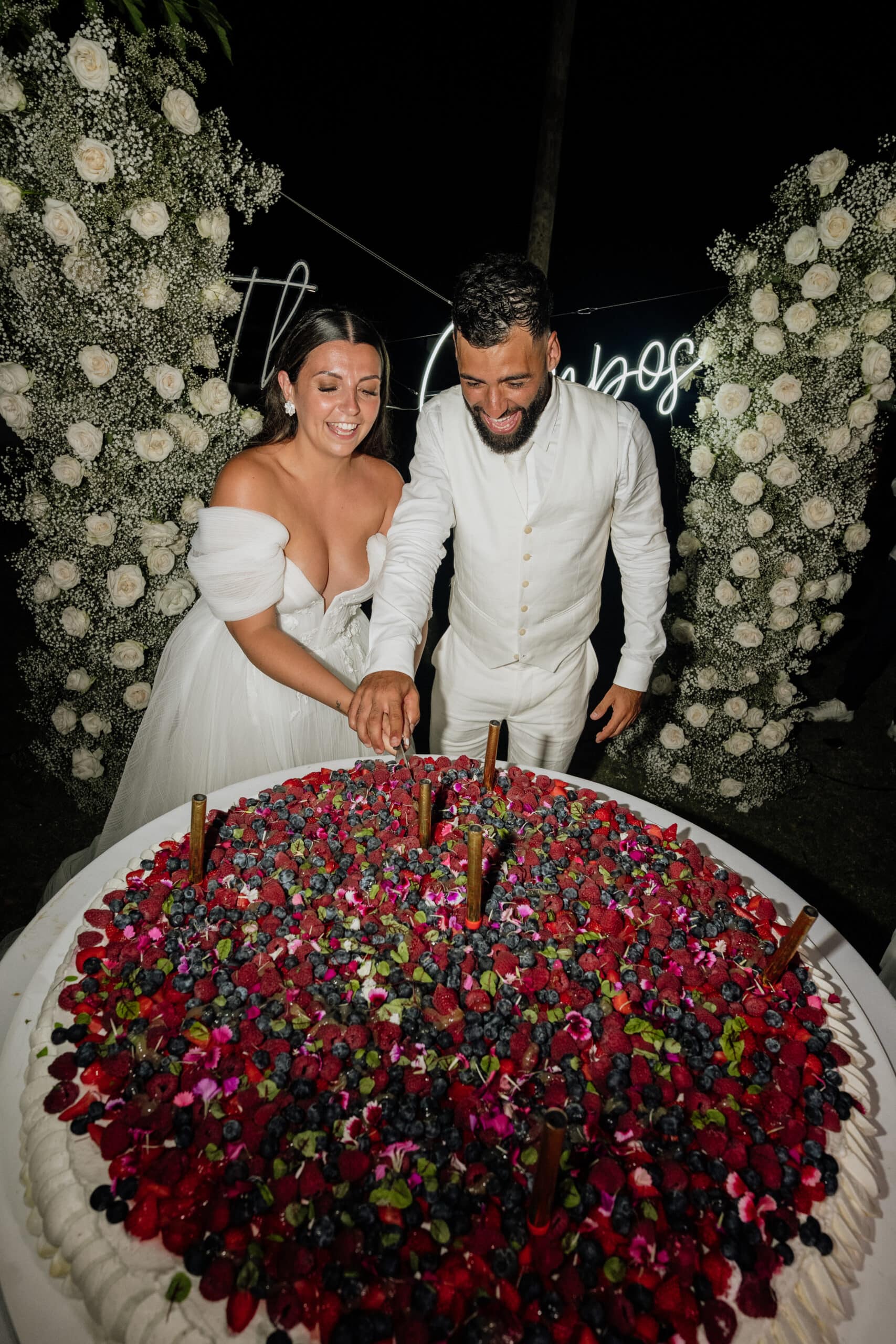 Jeunes mariés souriants coupant leur gâteau géant de mariage, richement décoré de fraises, framboises et myrtilles, devant des roses blanches.