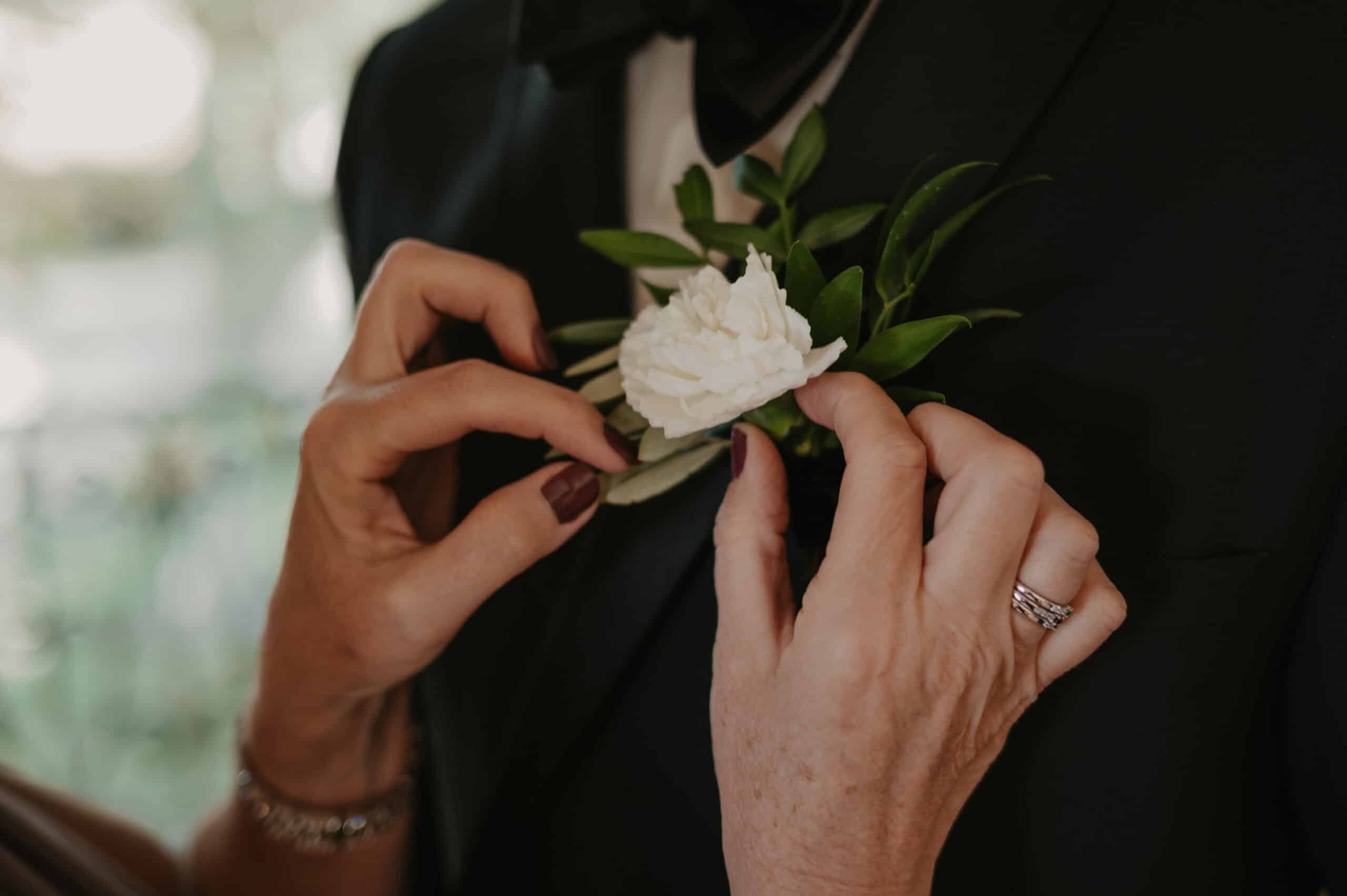 Des mains (avec des bagues et du vernis bordeaux) ajustent délicatement une boutonnière blanche sur le revers d'un costume noir de marié.