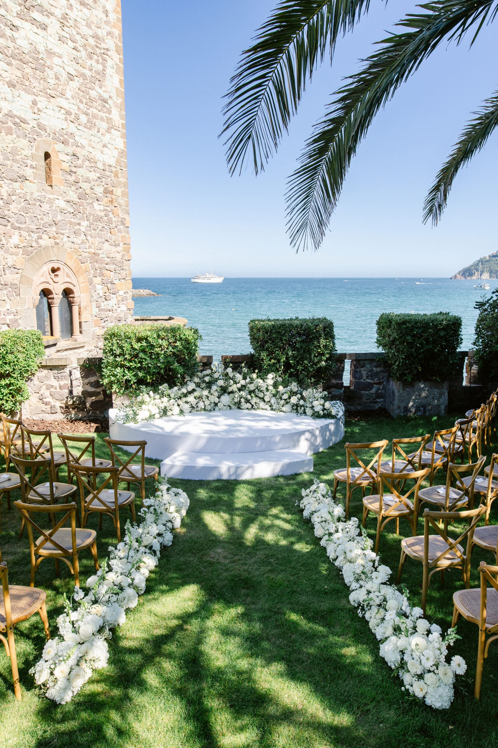 Élégante cérémonie de mariage extérieure sur pelouse. Autel blanc fleuri, chaises en bois et allée bordée de roses blanches, avec vue sur la mer.