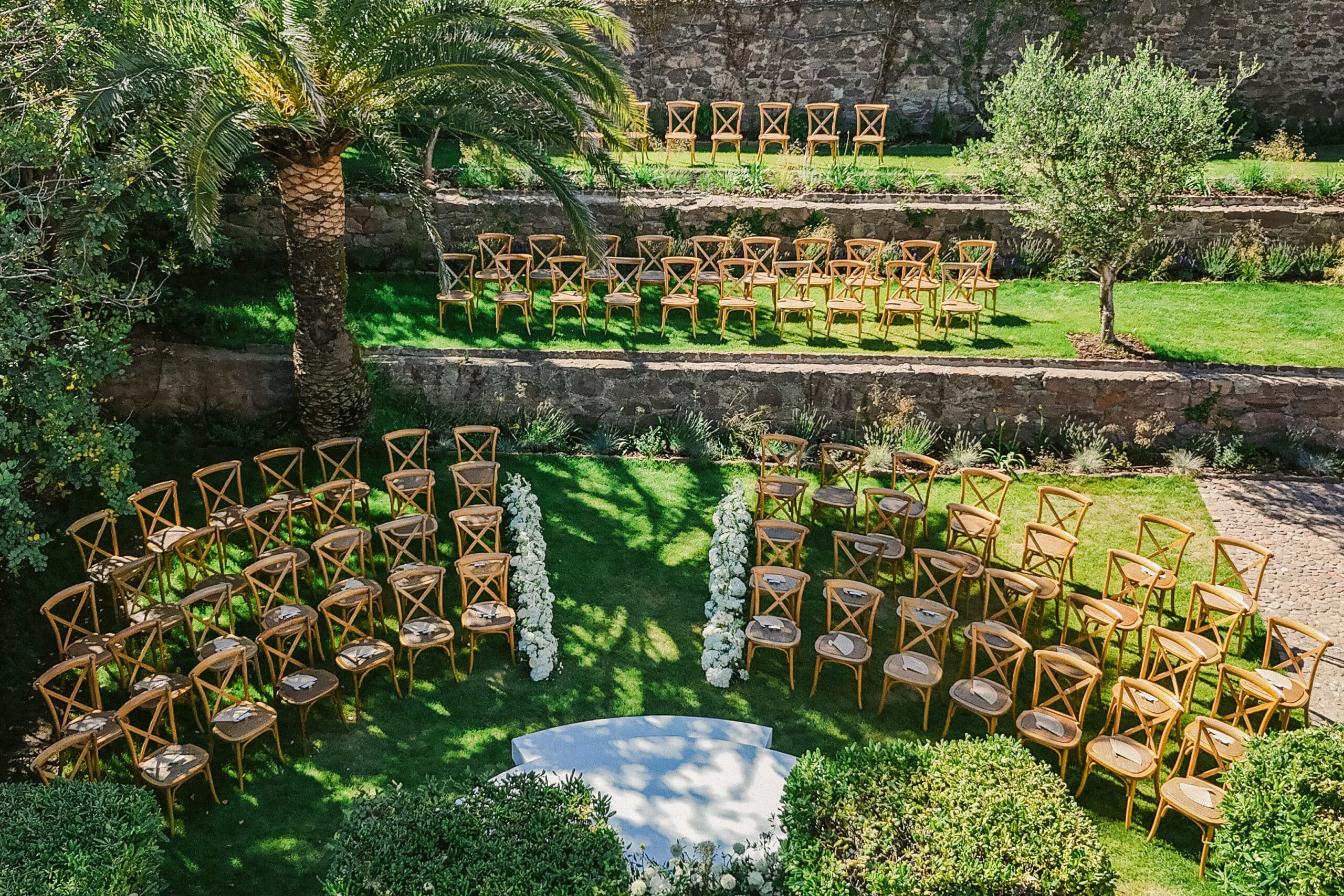 Cérémonie de mariage en plein air. Jardin en terrasses, chaises X-back en bois clair, allée fleurie et estrade blanche sous un palmier.