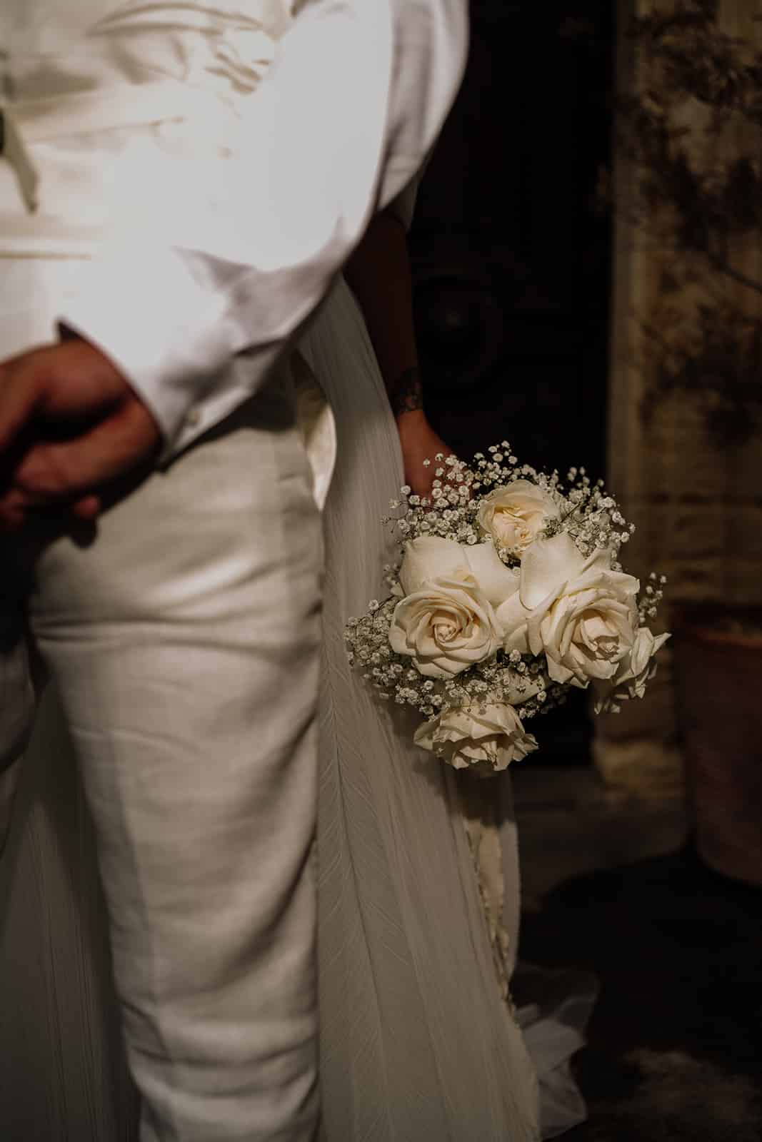 Bouquet de mariée intime de roses blanches et gypsophile. La mariée, main tatouée, et le marié en tenue claire se tiennent côte à côte.