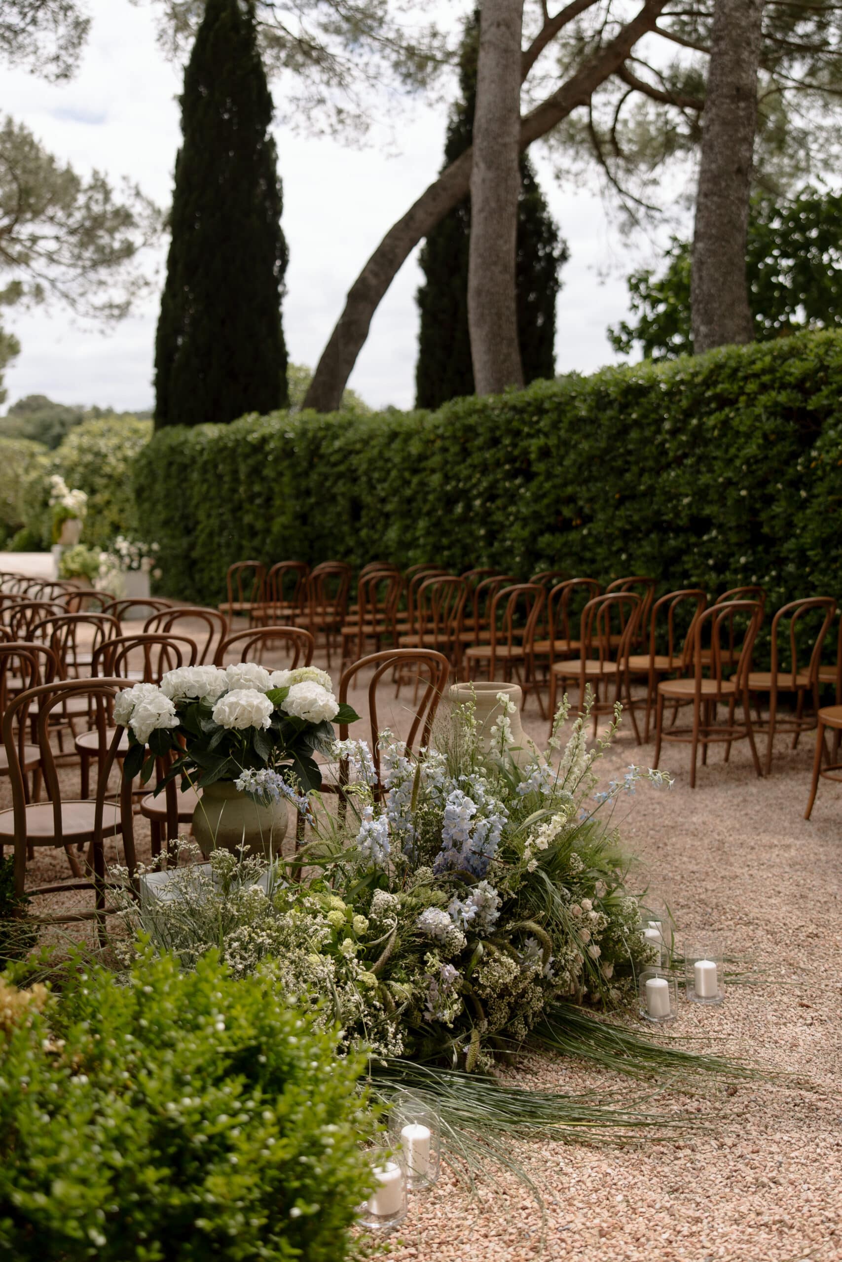 Allée de cérémonie de mariage en plein air bordée de chaises en bois courbé. Décoration florale sauvage (hydrangeas, delphiniums) sur un chemin de gravier.