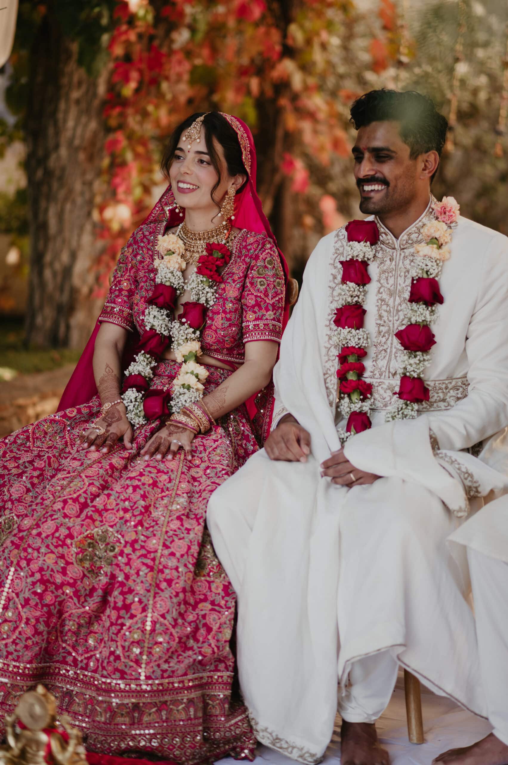 Couple indien souriant portant des guirlandes de fleurs, assis lors de leur mariage. La mariée est en lehenga rose vif brodé, le marié en sherwani blanc.