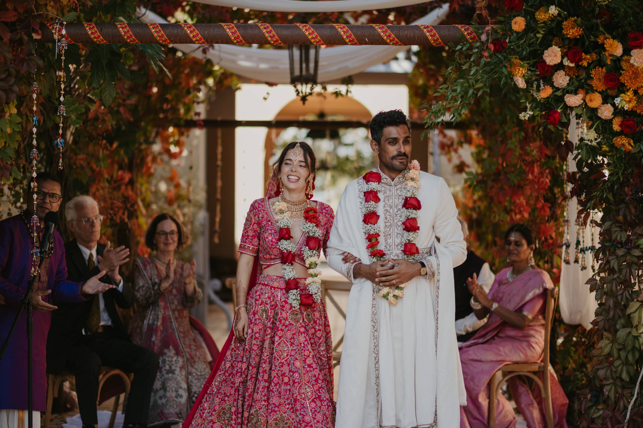 Mariée joyeuse en lehenga rose riant à côté du marié en sherwani blanc, sous un mandap décoré de fleurs et de vignes.