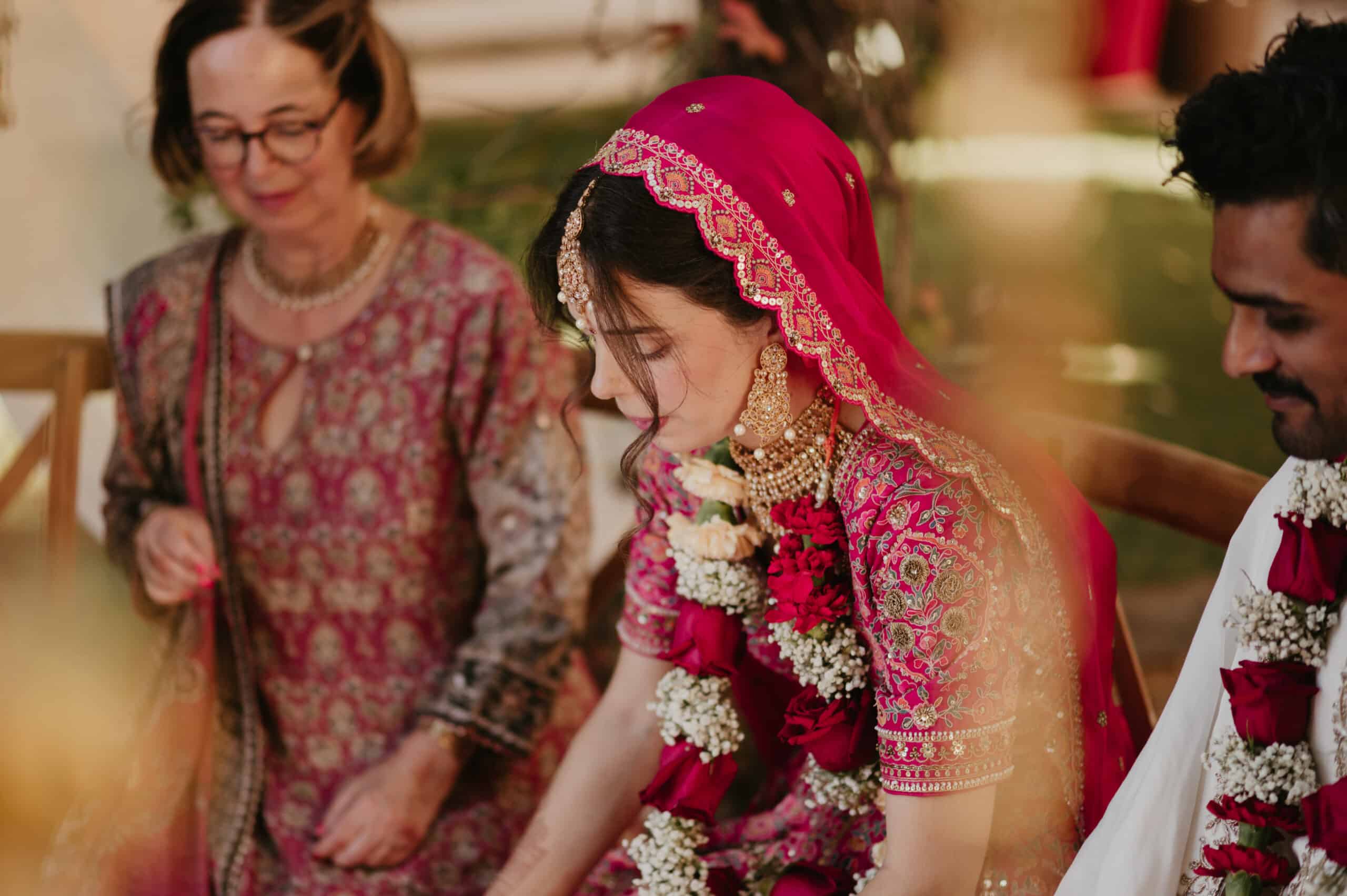 Mariée indienne portant une tenue rose fuchsia brodée et de somptueux bijoux traditionnels lors de la cérémonie, sa mère souriant à ses côtés.